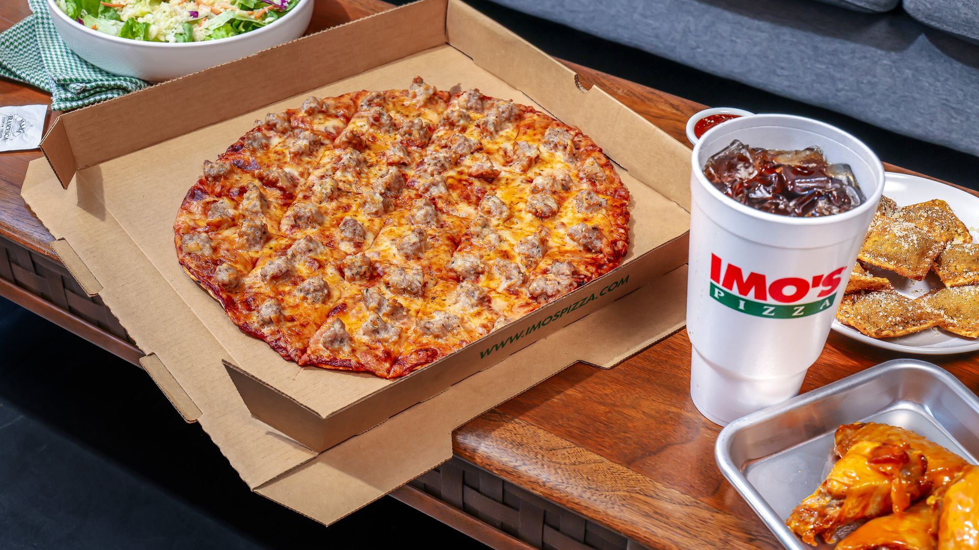 Large sausage-topped pizza in an open cardboard box on a wooden table, next to a bowl of mixed salad, a white soda cup, and a plate of breadsticks; a gray couch sits in the background.