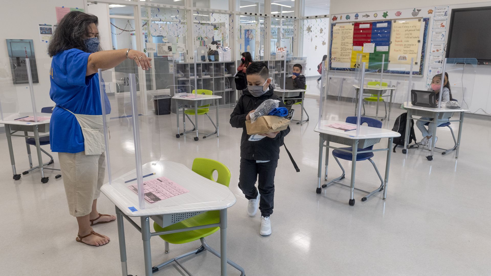 Picture of a classroom with desks six feet apart that have glass barriers, people are seen wearing masks
