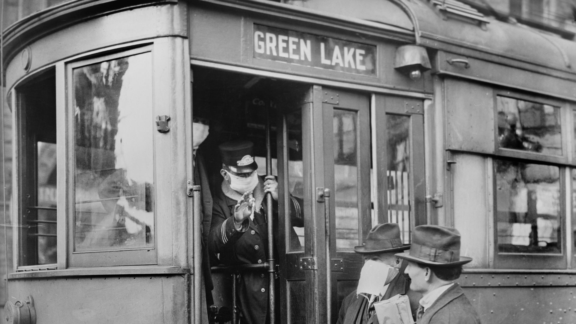 Two people stand next to a Seattle streetcar labeled "Green Lake" while a conductor signals to them, wearing a mask.