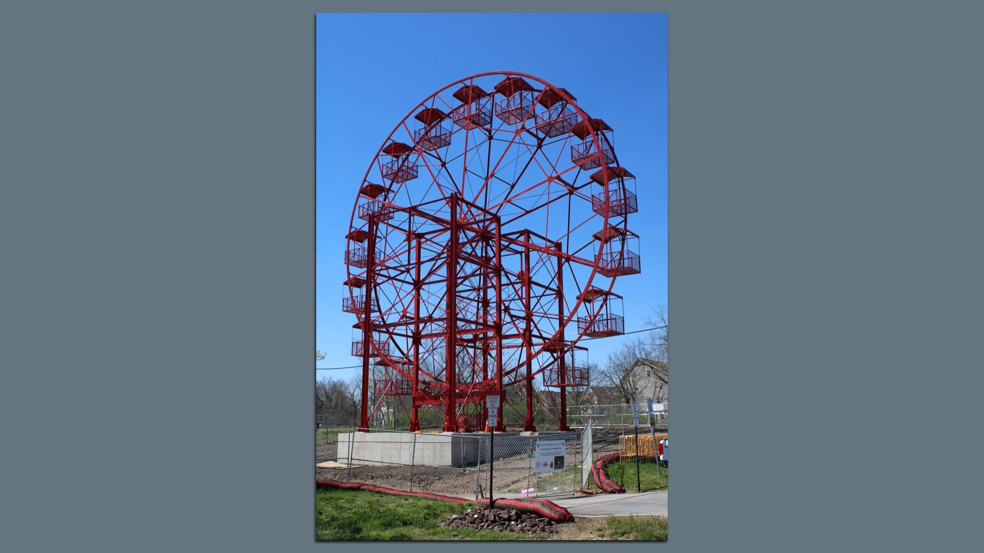 Red metal Ferris wheel with square cabins stands behind a chain-link fence in a park; a clear blue sky, supporting steel framework, and a few houses in the background.