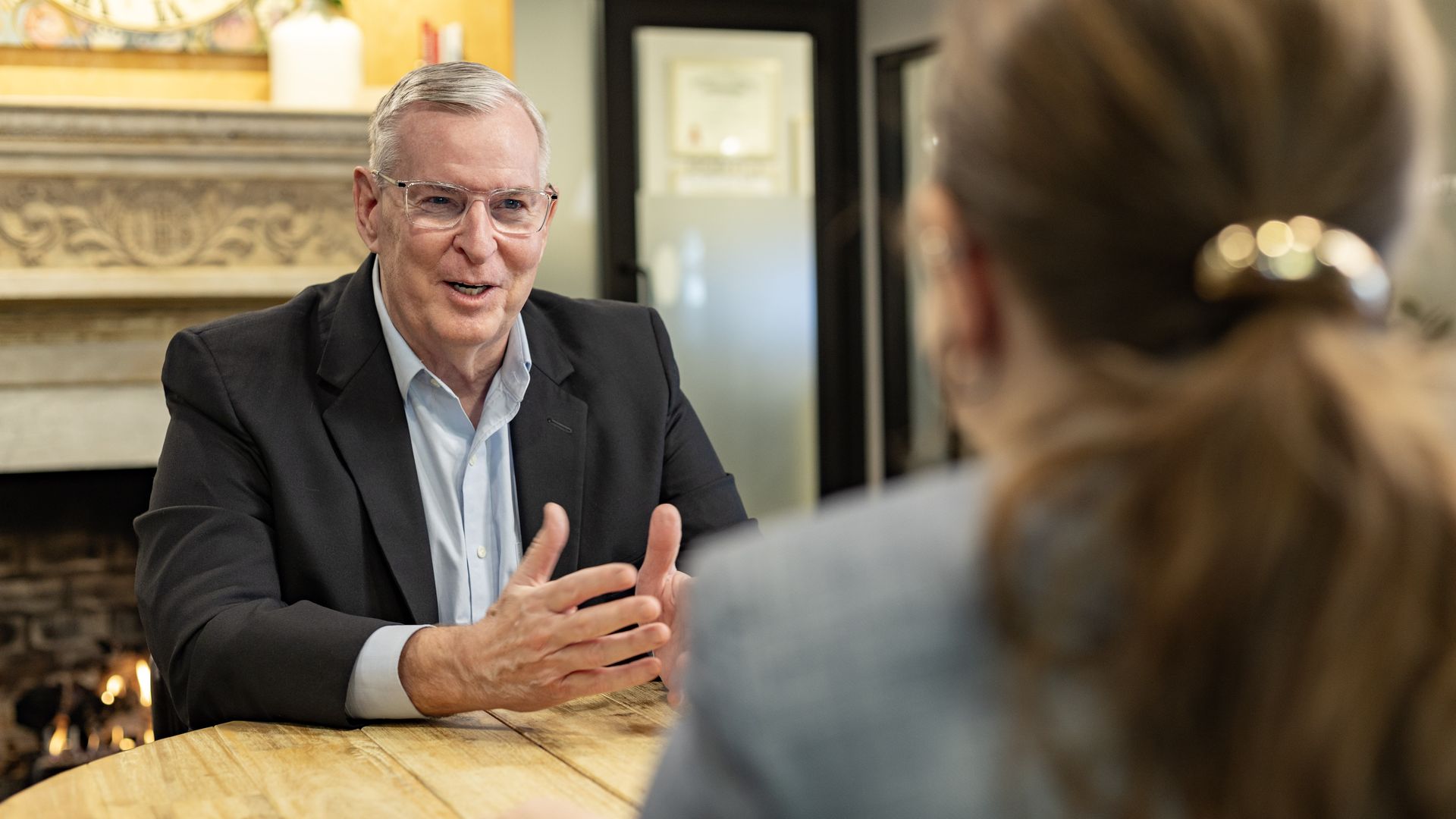 Older man in a black blazer and glasses speaks animatedly at a wooden table across from a woman with brown hair tied back, in a cozy room with a lit fireplace in the background.