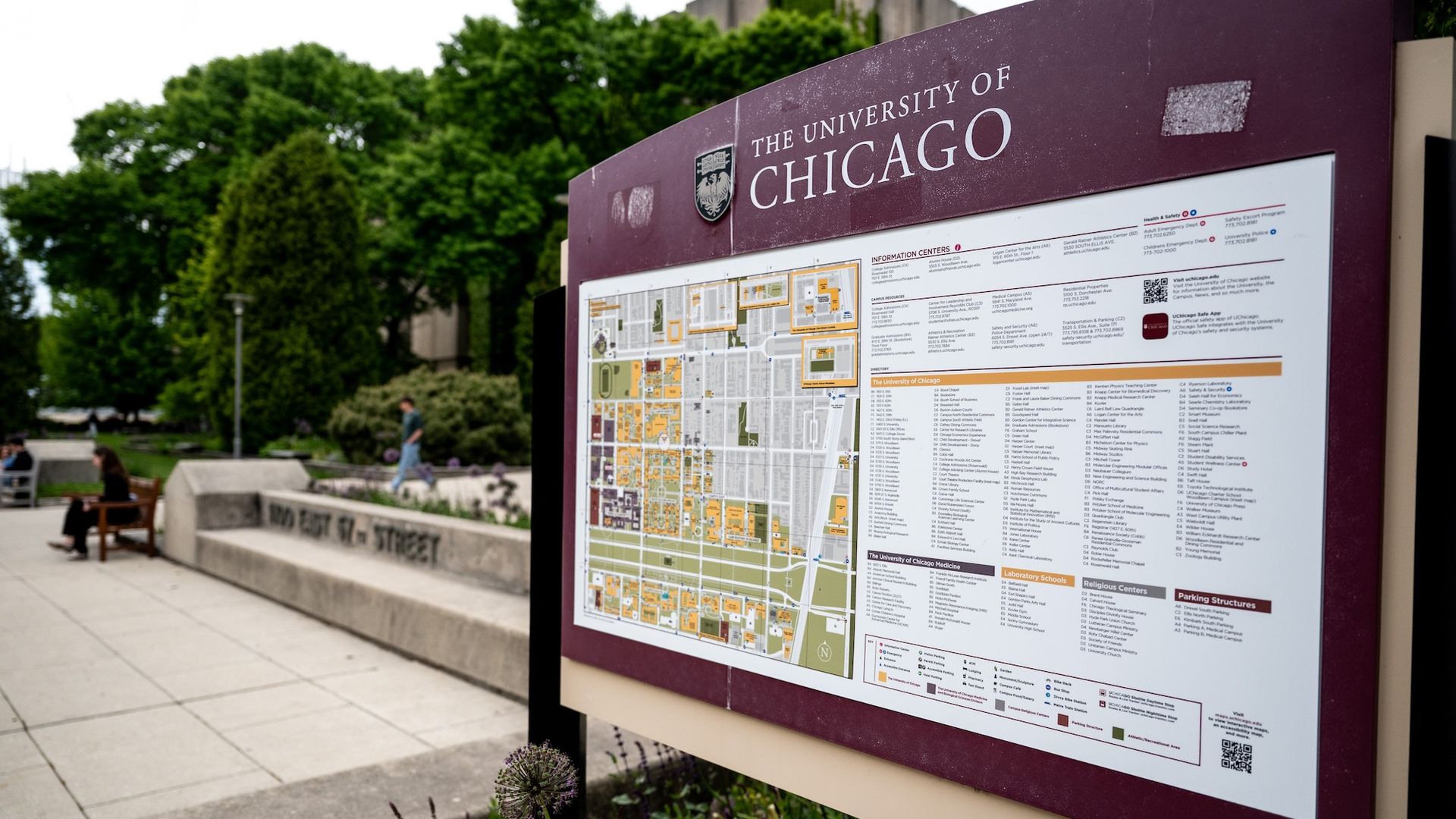 A large maroon campus sign reading "THE UNIVERSITY OF CHICAGO" sits beside a detailed map panel; green trees and benches in the background, with a person walking nearby.