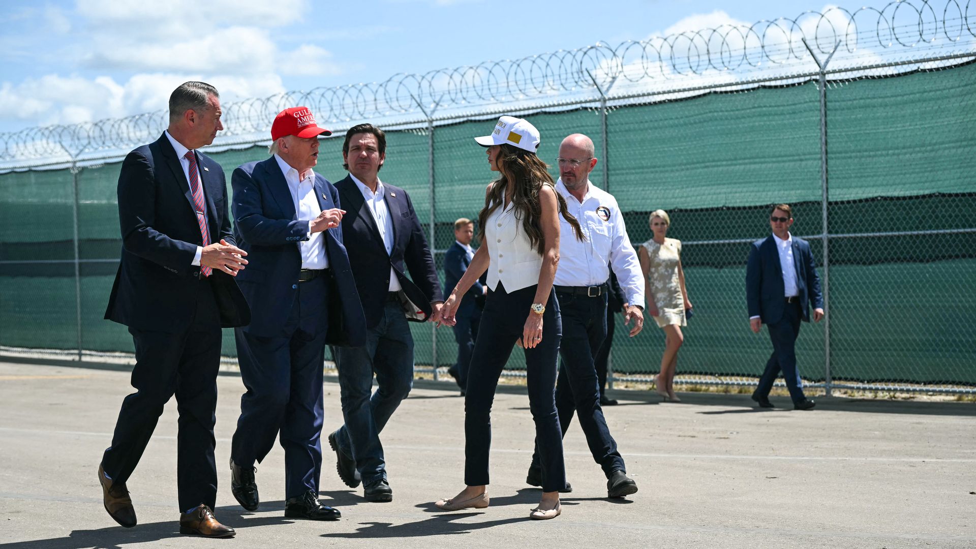 Six people, including President Trump, wearing a red hat, walk on a paved surface beside a high green fence topped with barbed wire under a blue sky with clouds.