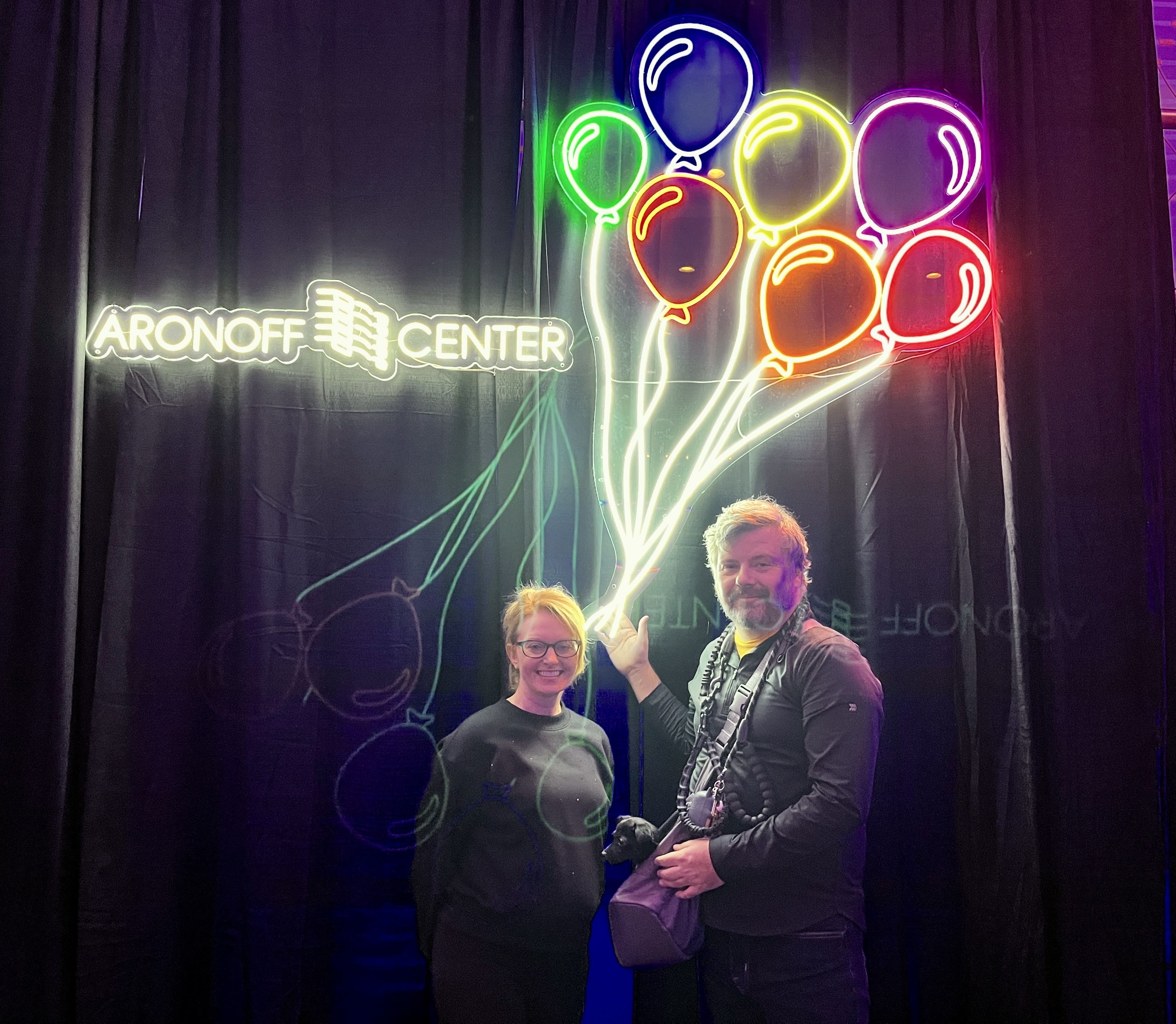 Bright neon balloon outlines in multiple colors on a dark backdrop with a white neon "ARONOFF CENTER" sign to the left; two people stand smiling in front.