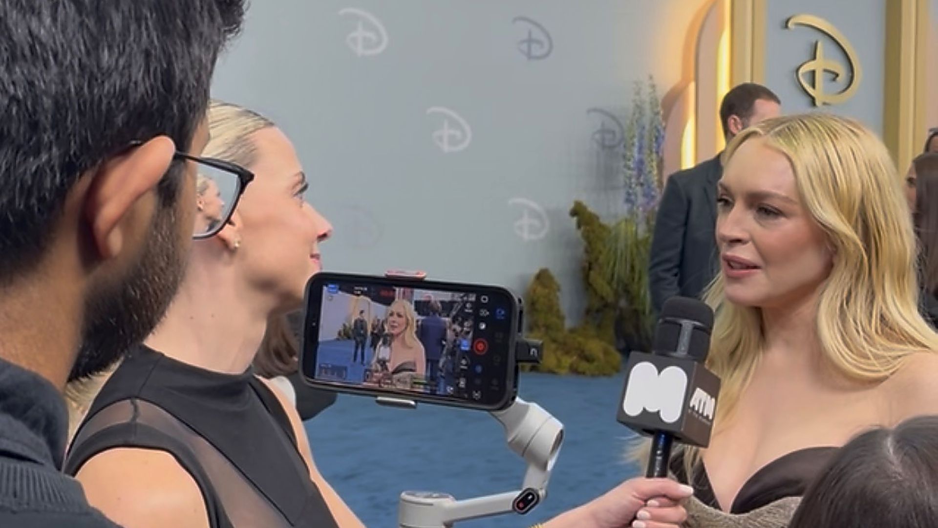 Blonde woman being interviewed speaks into a microphone as a man films with a smartphone on a stabilizer; blue backdrop with faint logos and a doorway to the right, others nearby.