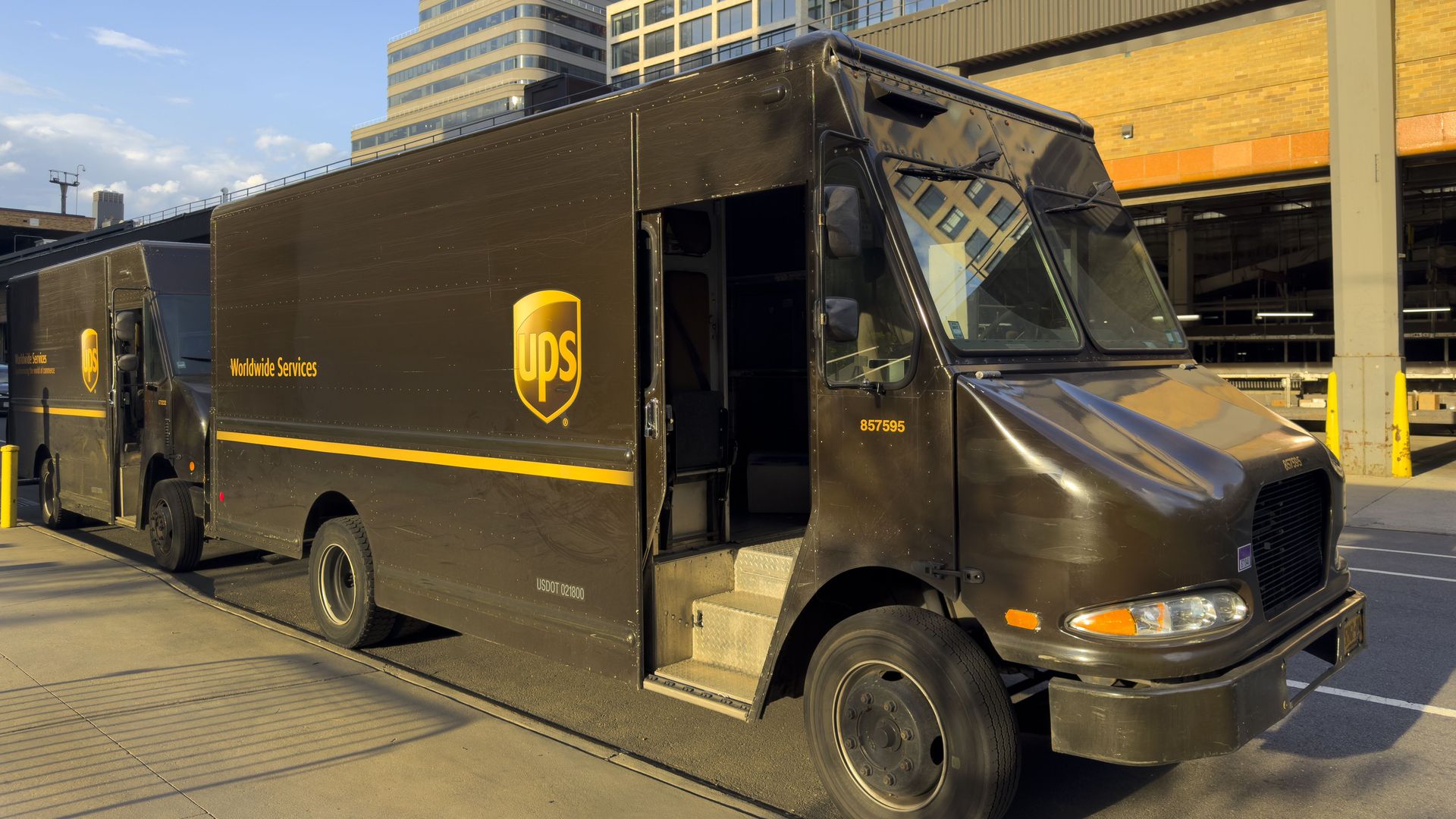 Two brown UPS delivery trucks parked outside a building with open driver-side doors, yellow UPS logo and 'Worldwide Services' text visible, under a clear blue sky.