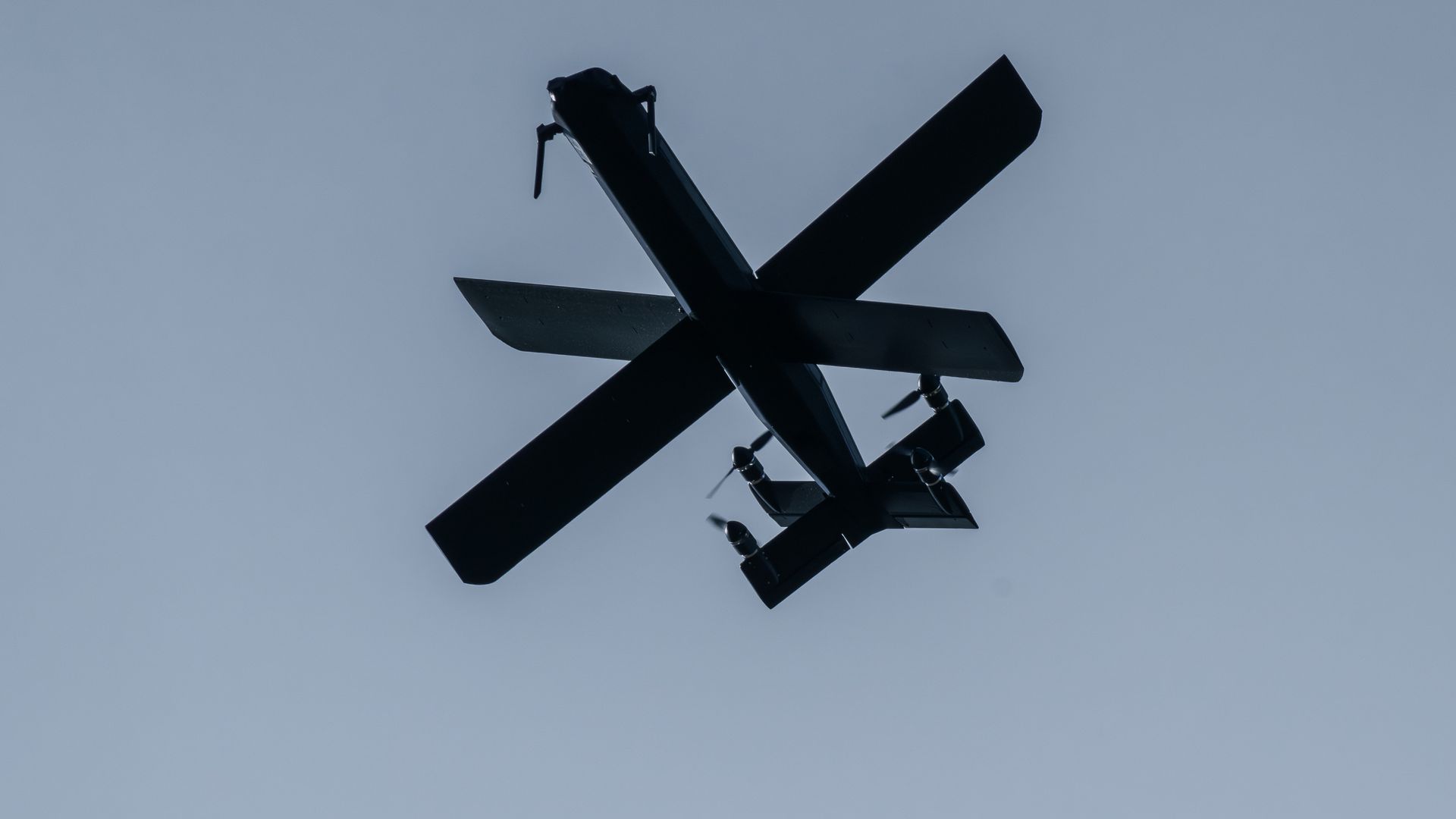 Silhouette of a drone with large blades crossing in an X shape. Set against a dark blue sky.