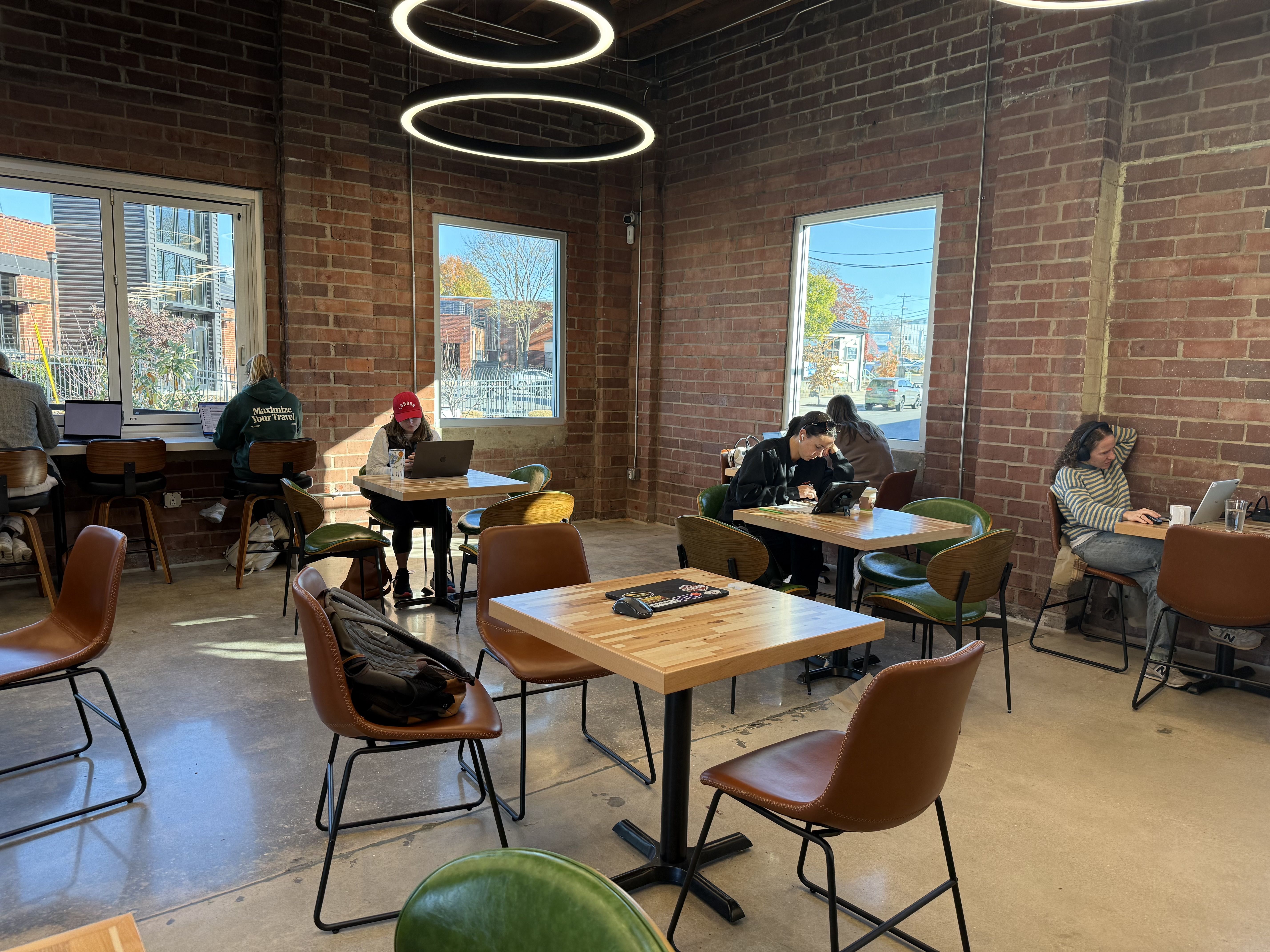 Cozy cafe with brick walls and large windows showing fall trees outside. People working on laptops at wooden tables and leather chairs under circular ceiling lights.