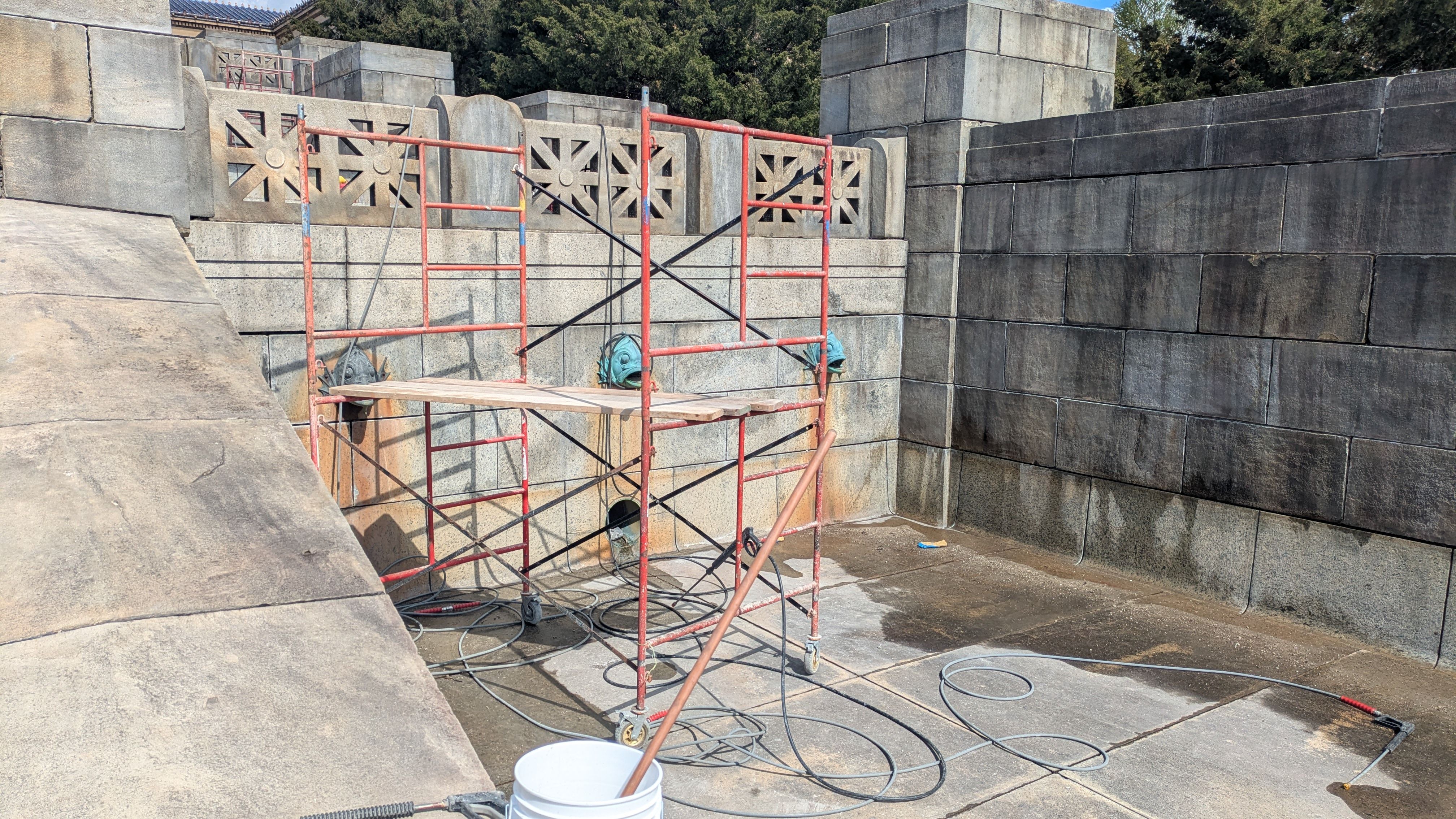 Red scaffolding sits in a stone courtyard with large gray block walls and decorative cutouts. Hoses, a white bucket, and a long pipe rest on the wet concrete floor.
