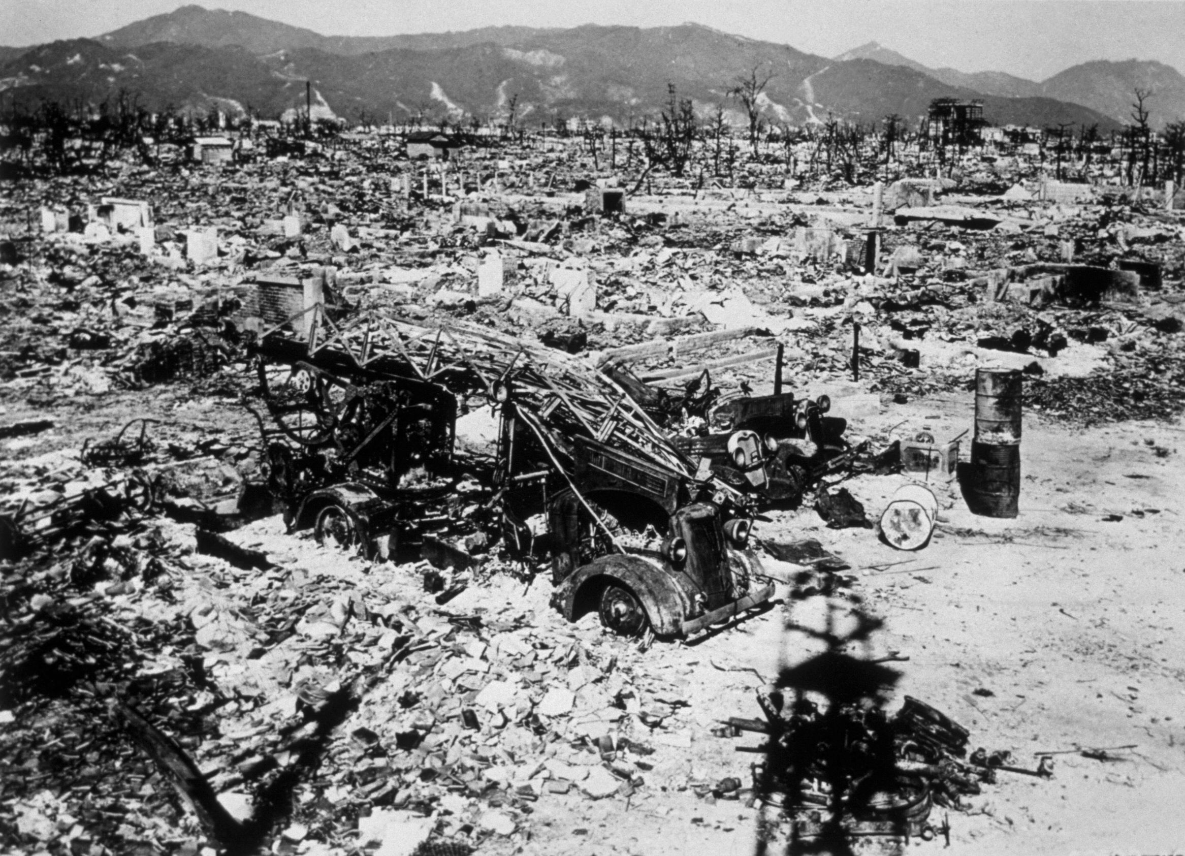 Black and white photo showing destroyed vehicles and ruins scattered across a vast area with damaged trees and mountains in the background.