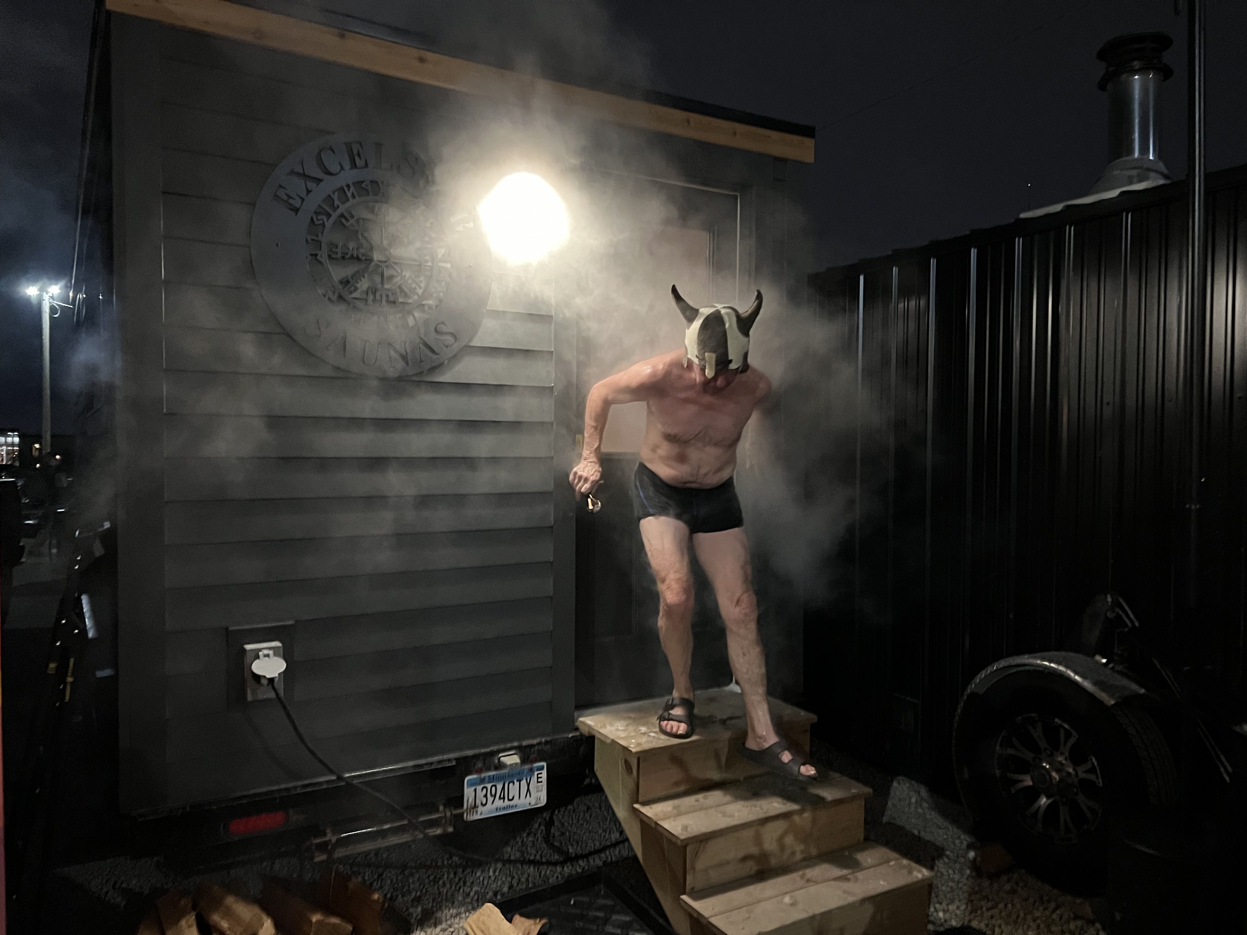 A man in a bathing suit, sandals and a viking hat emerges in a cloud of steam from a dark mobile sauna building.