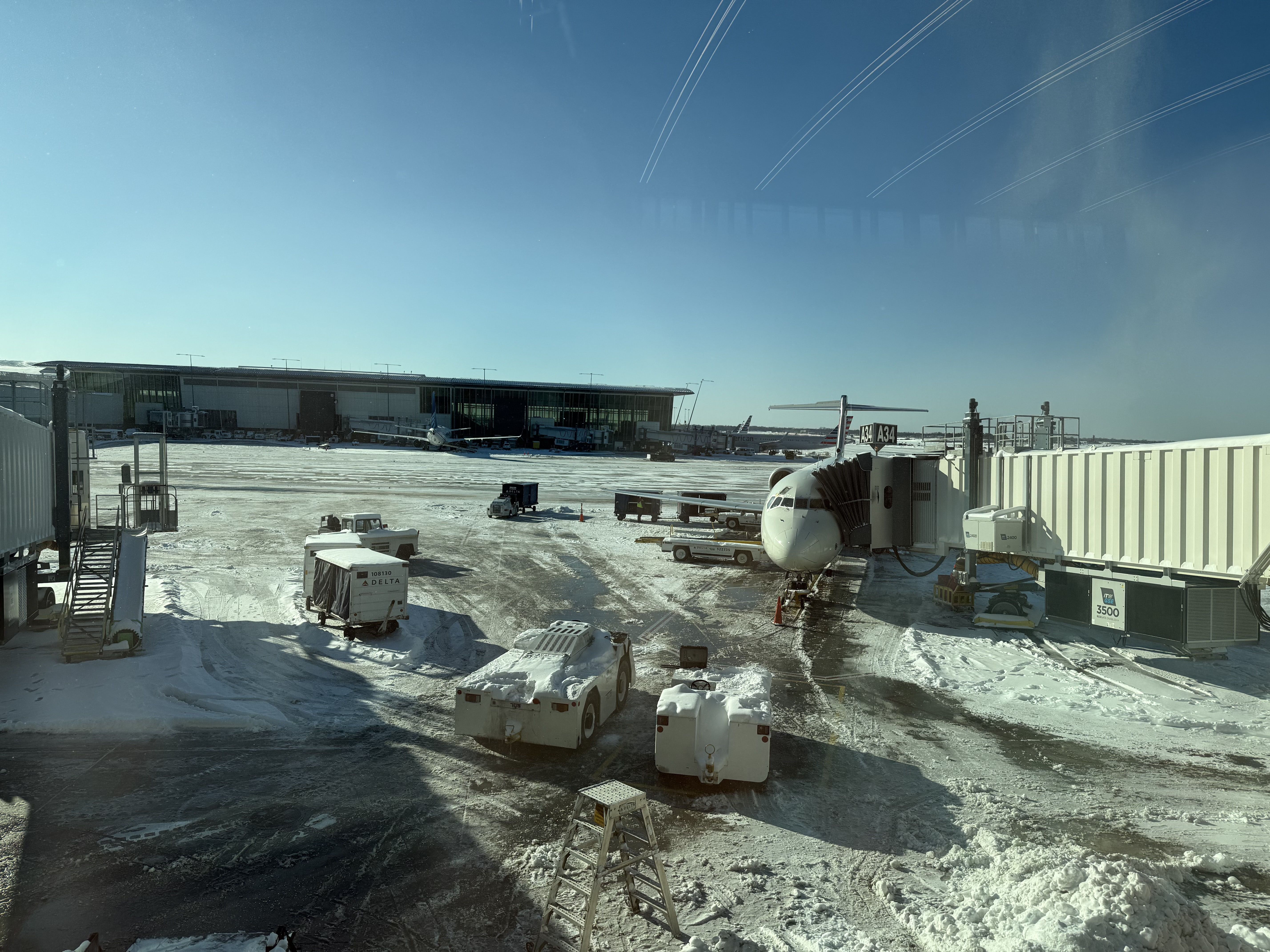 Snow-covered airport tarmac with a plane docked at gate A34, ground service vehicles, equipment, and a clear blue sky overhead.