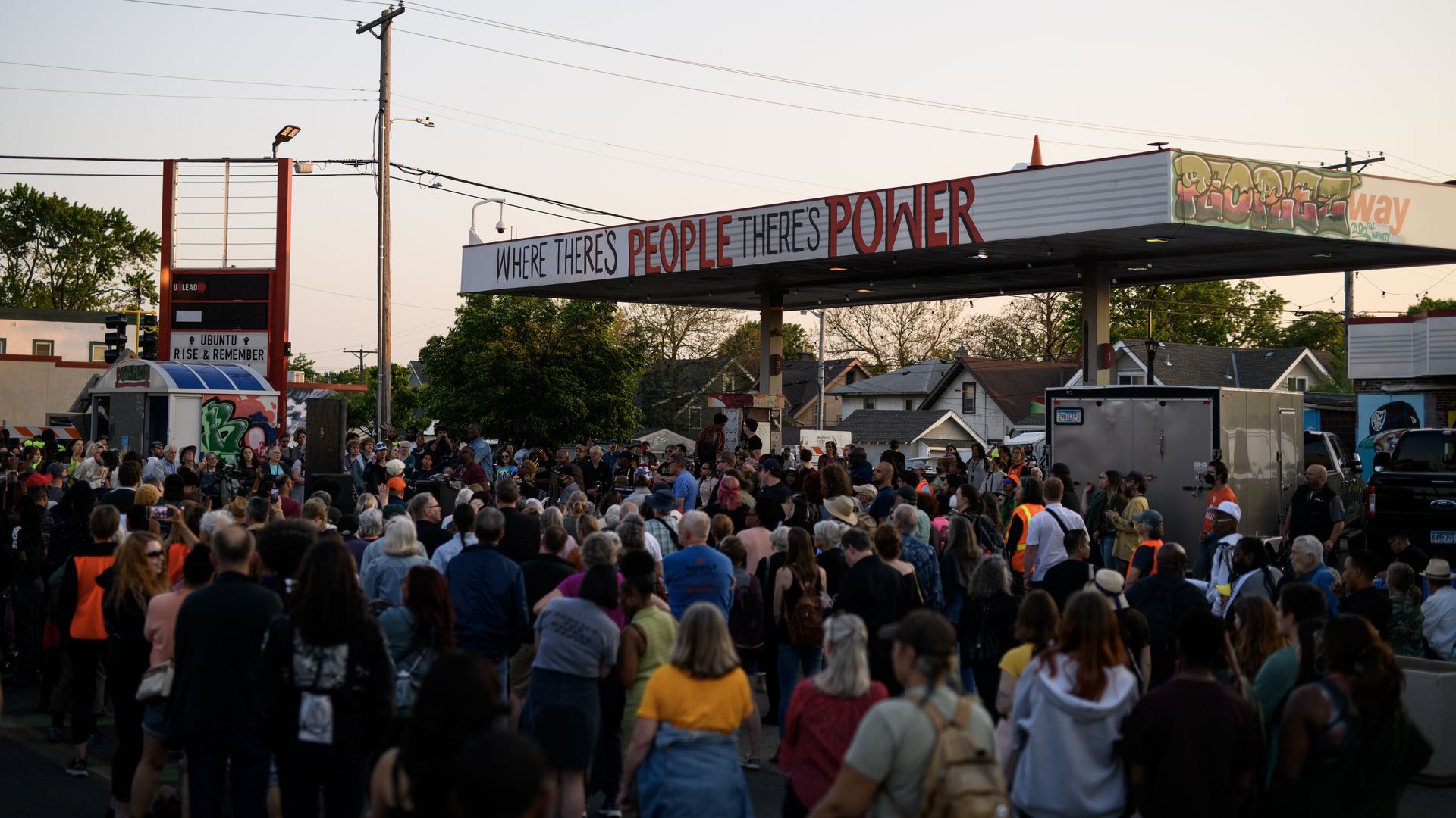 A large group of people gather in George Floyd Square with the former gas station - with the inscription "Where There's People There's Power" - in the background