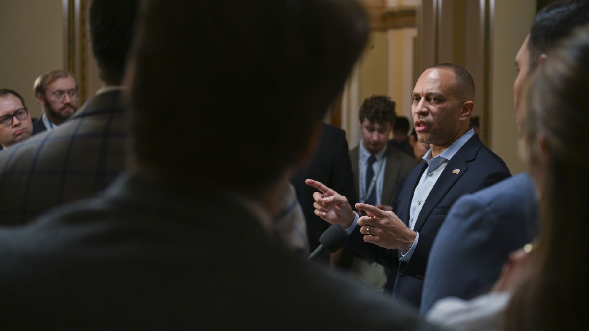 Hakeem Jeffries, wearing a blue suit with no tie, speaks to a gaggle of reporters in a beige hallway.