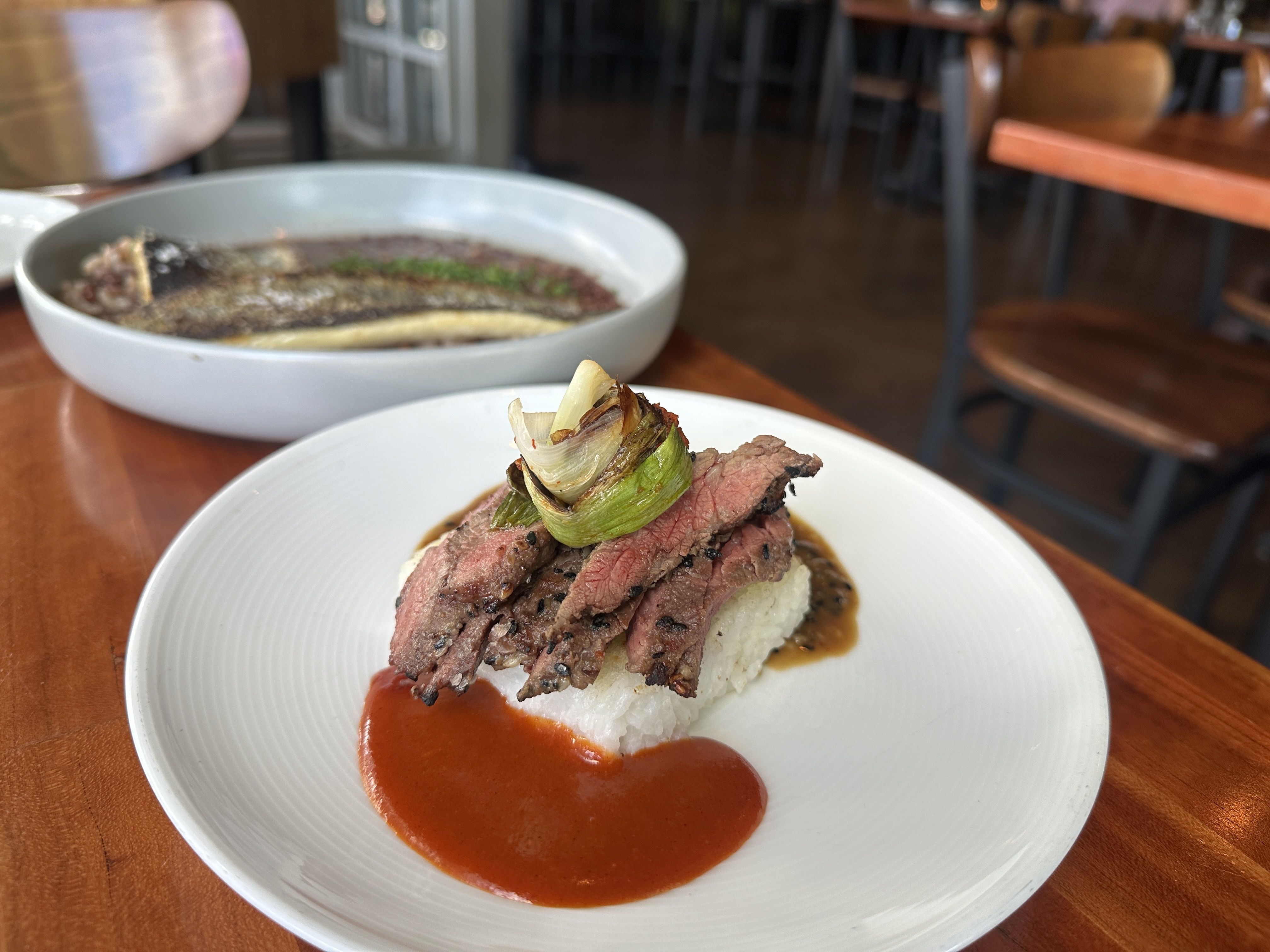 Plated dish with sliced steak and grilled green onion on rice, surrounded by sauces on a plate, with a bowl of cooked fish in broth in the background at a wooden table.