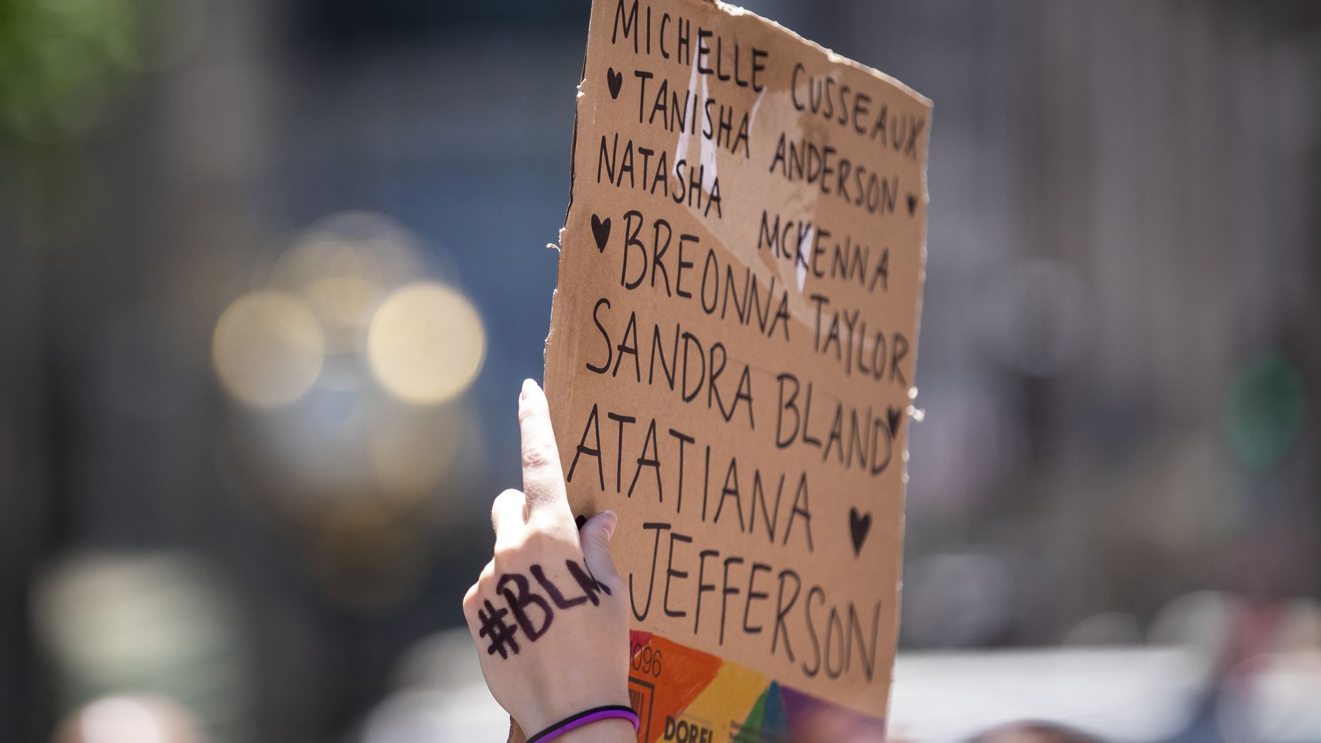 A protester holds up a homemade sign on a box that says, "Breonna Taylor, Atatiana Jefferson, Sandra Bland Natasha McKenna, Tanisha Anderson, Michelle Cusseausx" with the letters "#BLM" written on their hand and the rainbow colors on the box