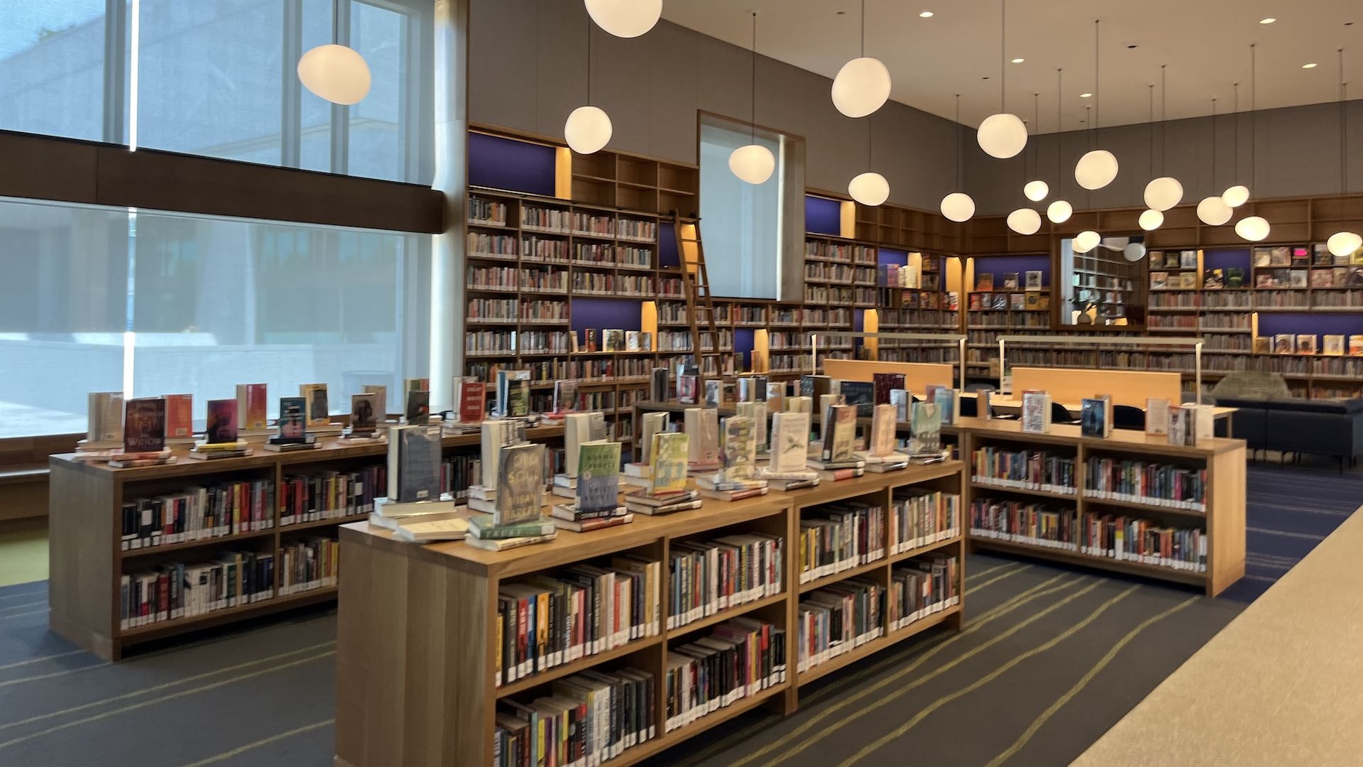 Modern library interior with tall windows on the left, rows of wooden bookshelves, and numerous round pendant lights hanging from the ceiling. Foreground shows tables displaying books.