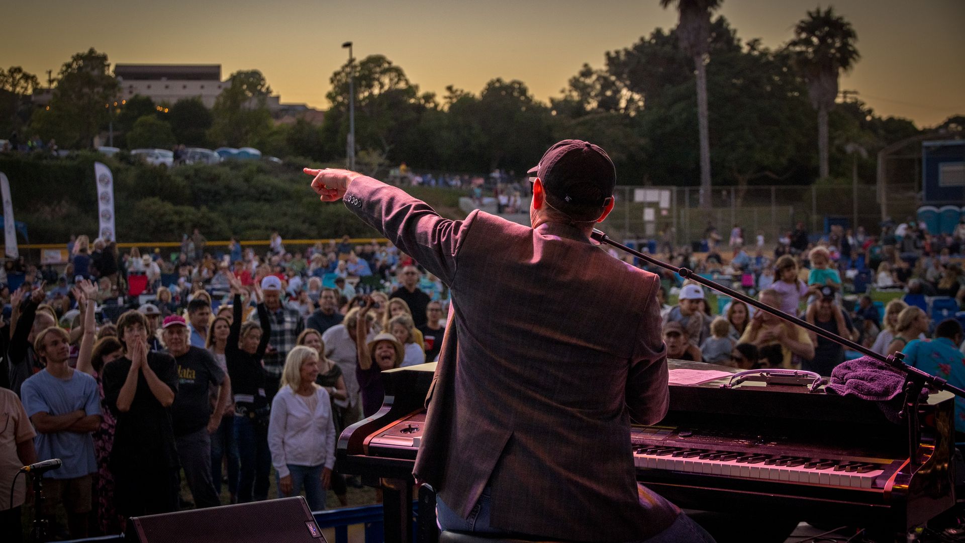 Back view of a musician in a blazer and cap at a keyboard on an outdoor stage, pointing toward a cheering crowd as sunset lights the sky with an orange glow behind trees.