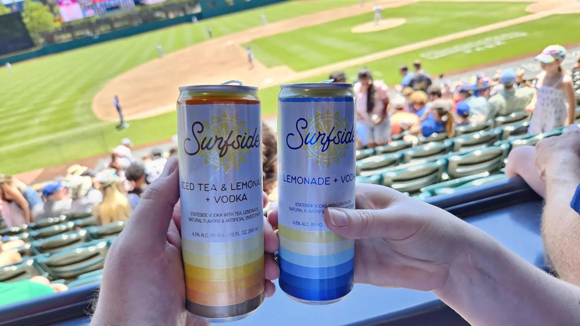 Hands hold up two "Surfside" lemonade + vodka cans with a baseball game played in the background.