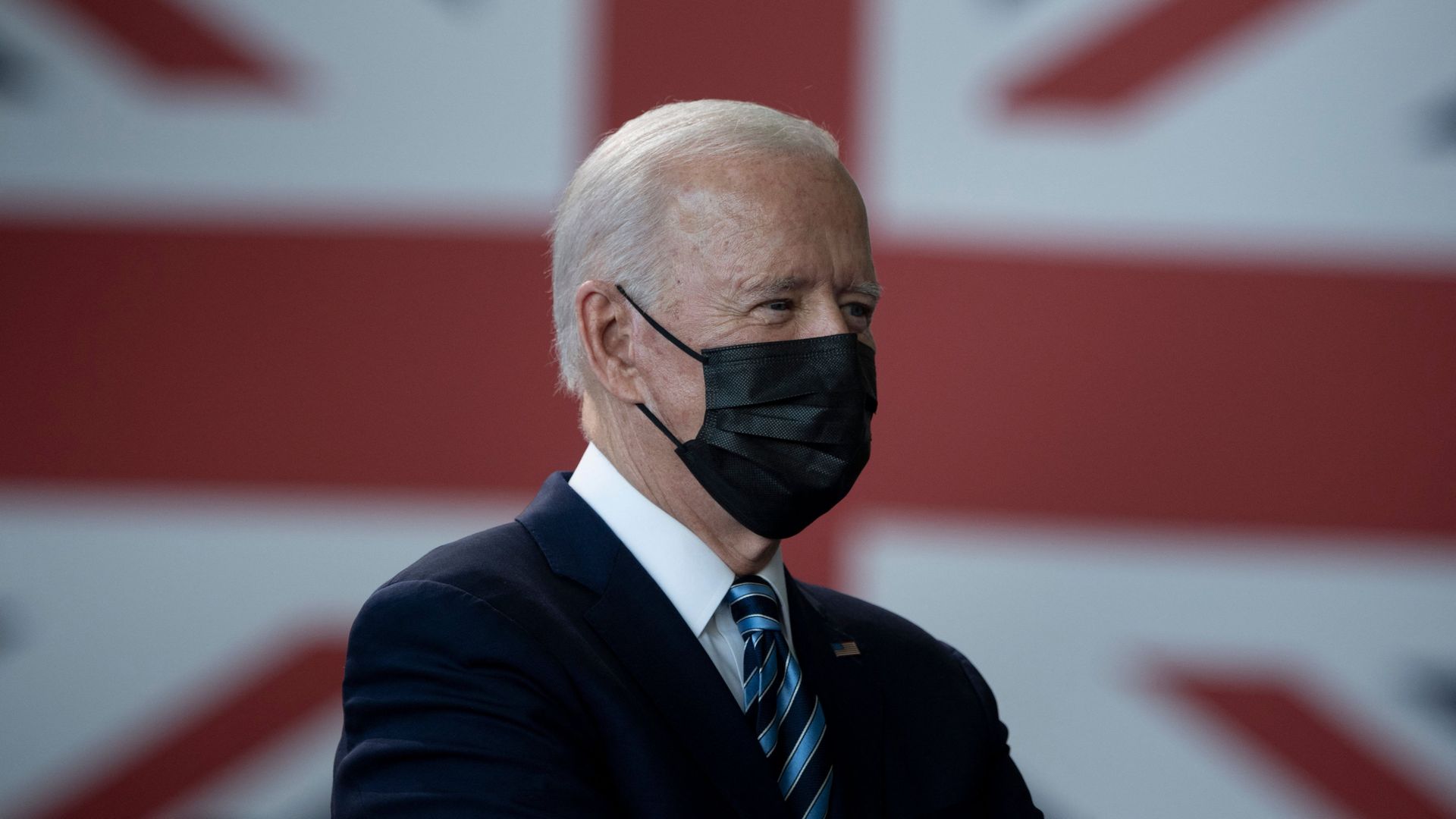 Picture of President Biden wearing a mask while standing In front of a British flag