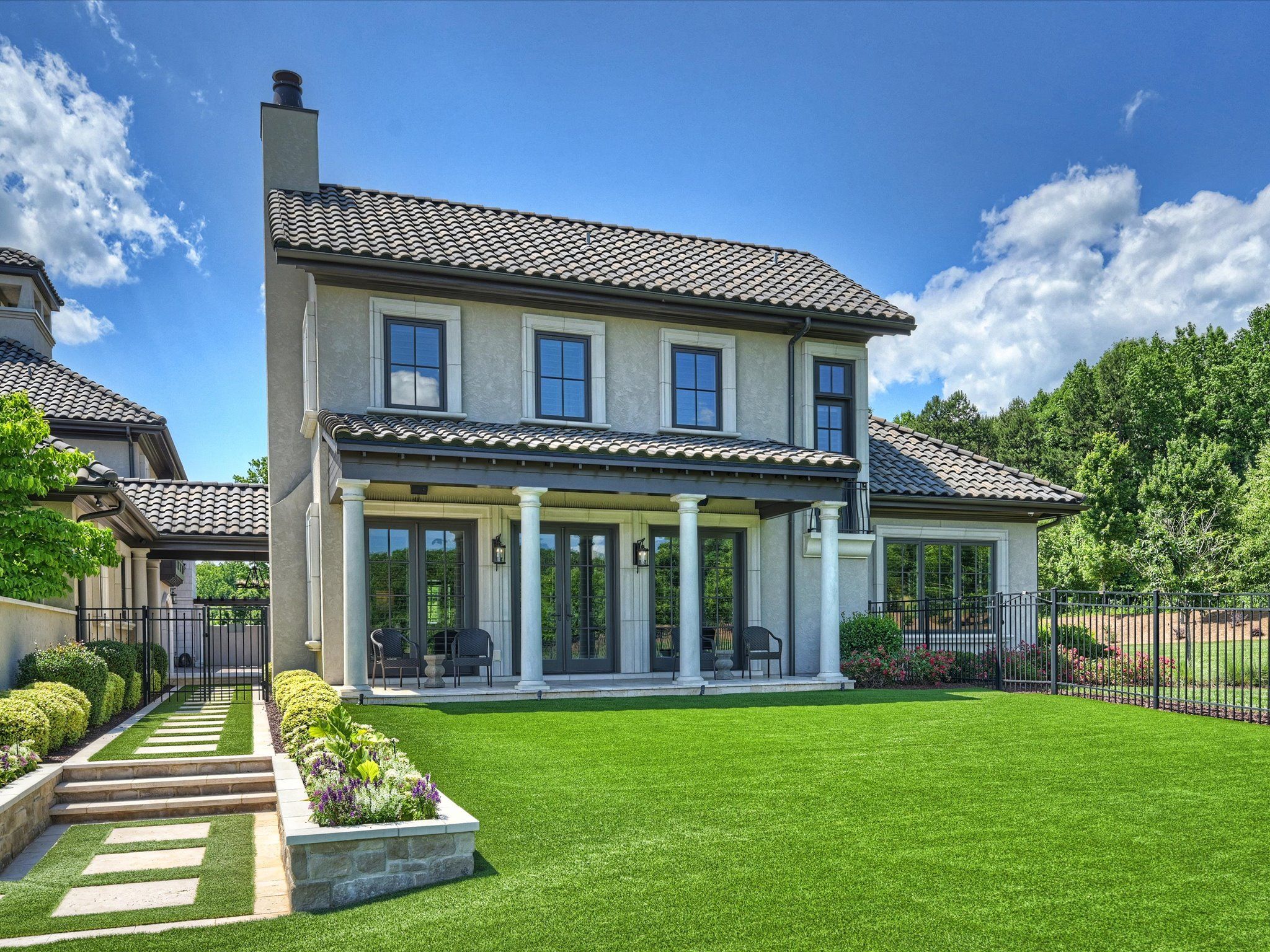 Two-story beige house with tiled roof, white pillars, and black-framed windows, surrounded by a manicured green lawn, flower beds, and a black metal fence under a blue sky with clouds.