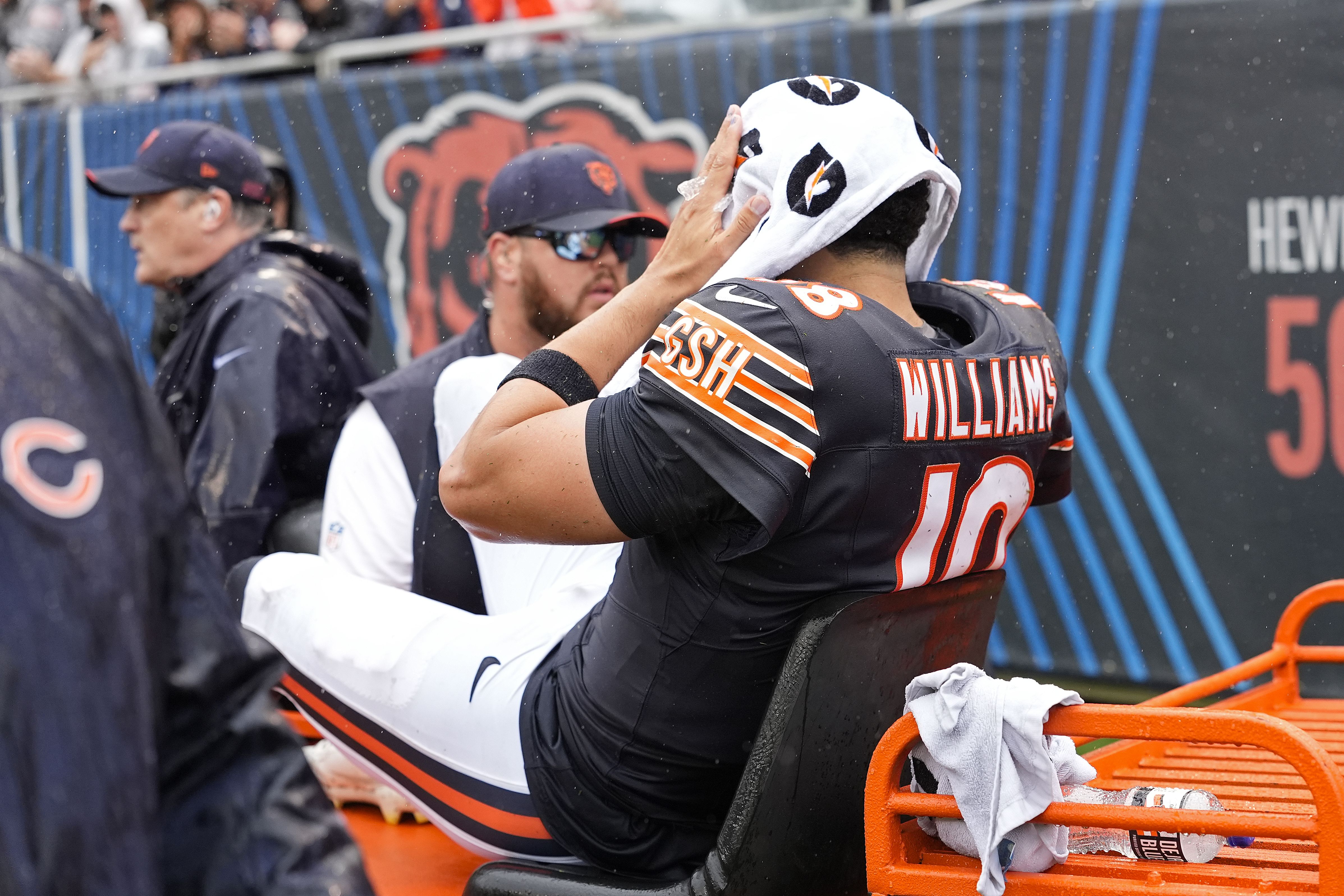 Chicago Bears football player wearing #10 Williams in a dark jersey with orange trim sits with a white towel on his head as rain falls; teammates and a stadium backdrop are visible.