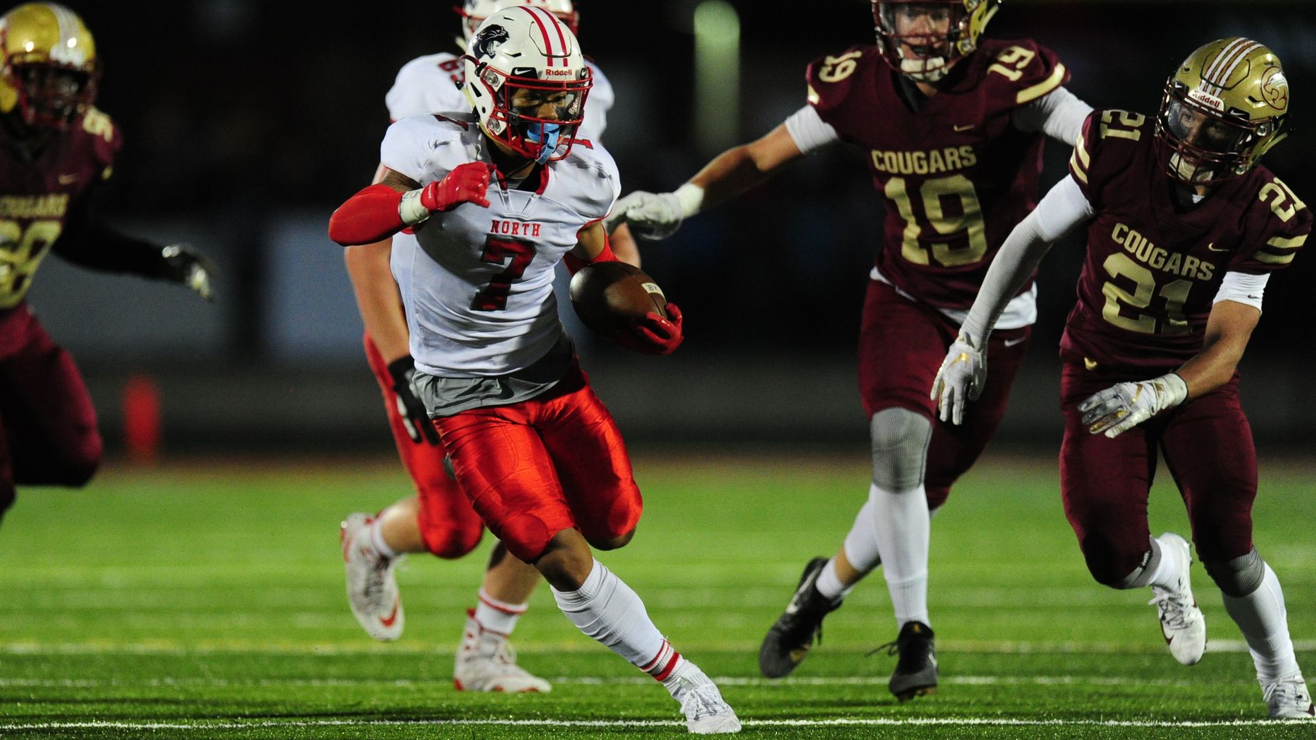 A football running back in a white jersey, white helmet and red pants cuts to his right as defenders wearing maroon uniforms and gold helmets chase him 