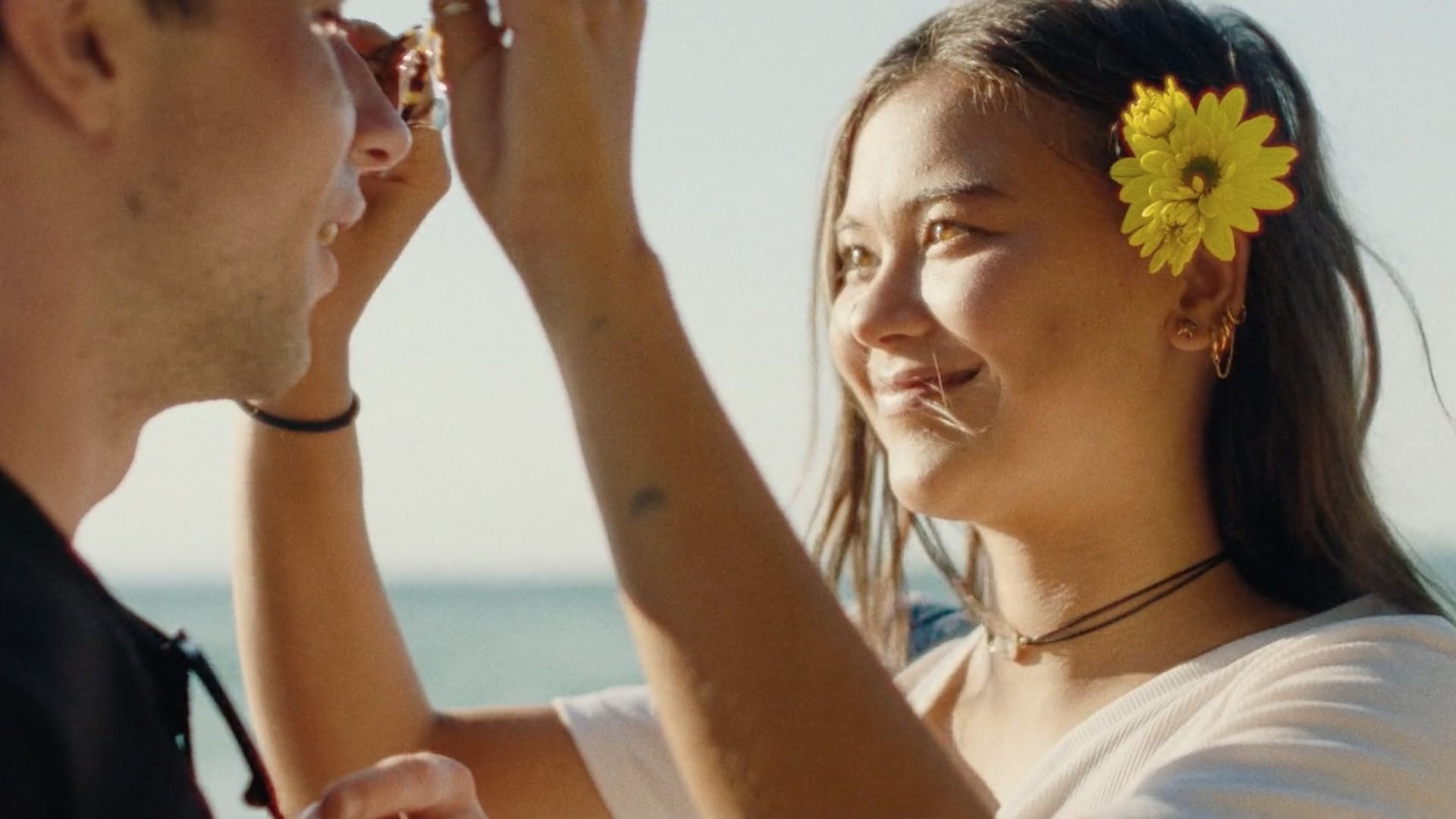 Sunlit beach moment: a woman with a yellow flower in her hair smiles at someone in sunglasses; she wears a white top and gold jewelry, with the sea in the background.