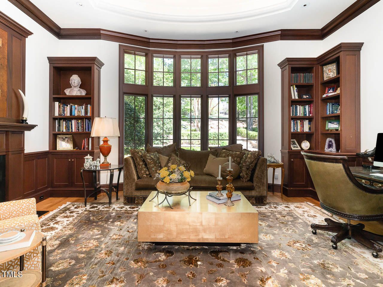 Elegant living room with large wooden windows showing greenery outside, brown sofa with pillows, gold coffee table with flowers and candles, wood bookshelves, and patterned rug.