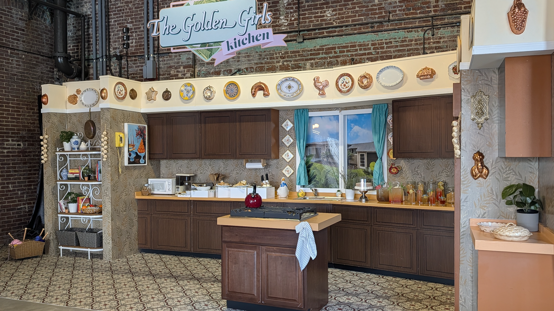 Vintage-style kitchen set with dark wood cabinets, patterned floor tiles, turquoise curtains, decorative plates on wall, and a sign reading "The Golden Girls kitchen" above.