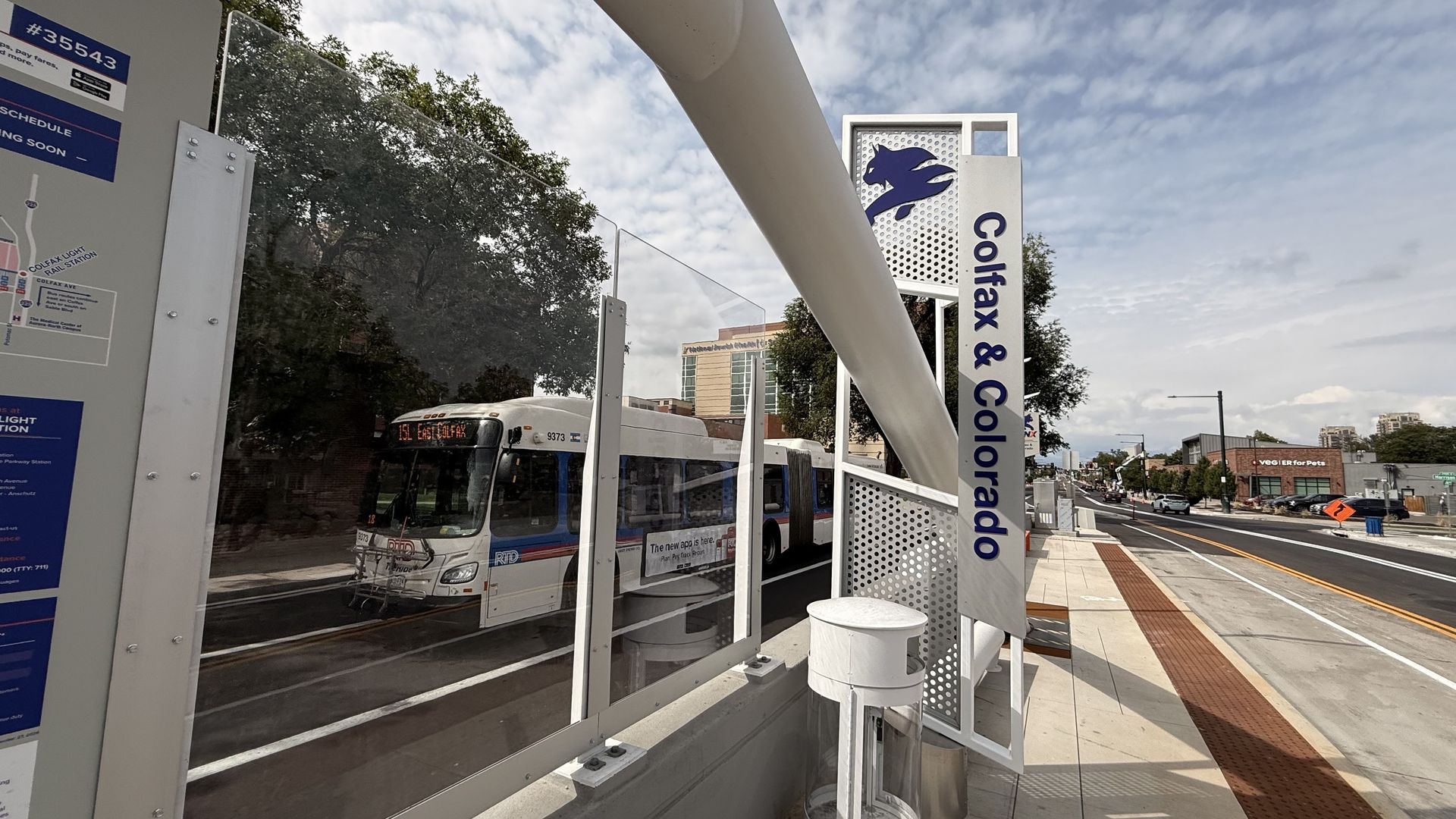 Street view at Colfax & Colorado, showing a modern bus stop with glass panels, perforated metal sign with a blue animal logo and city bus at the curb, and a blue-sky, cloud-filled city street.