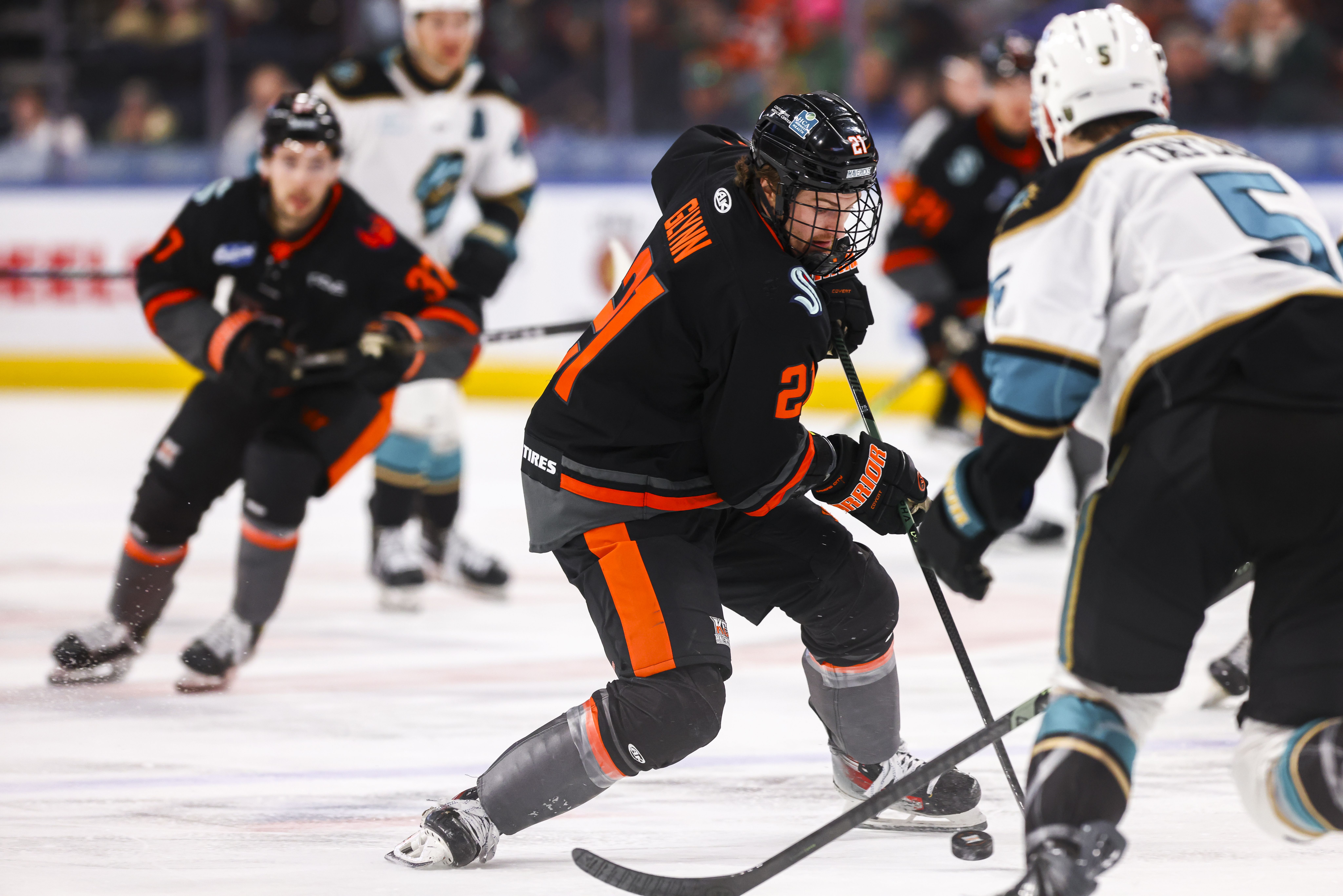 Hockey player in Mavericks black and orange jersey with number 27 controlling puck on ice, facing off against player in white and teal jersey with number 5, with blurred players in background.