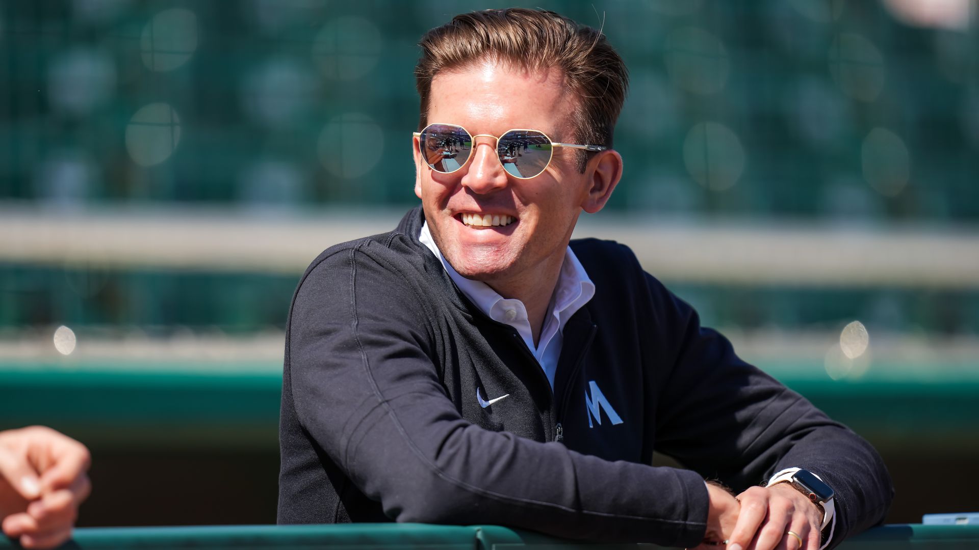 A smiling man with brown hair, light skin and tinted sunglasses leans over a bullpen railing on a baseball field