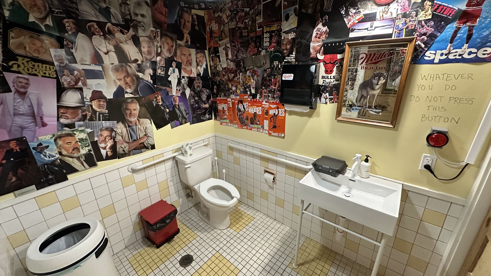 A wide-angle view of a white and yellow tiled bathroom with walls covered in posters of Kenny Rogers and Michael Jordan.