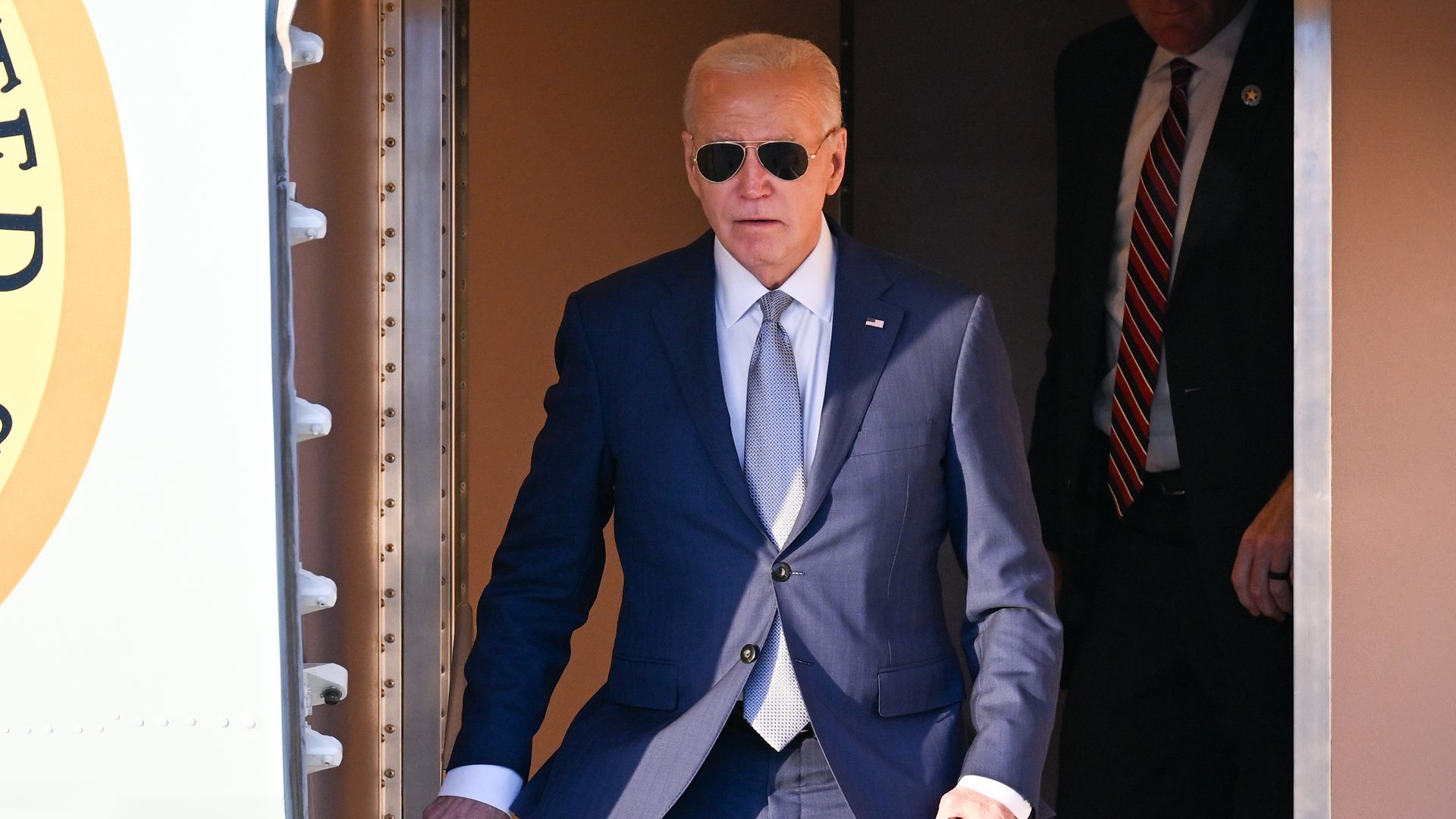 President Biden, wearing a blue suit, aviator sunglasses and stepping out of Air Force One.