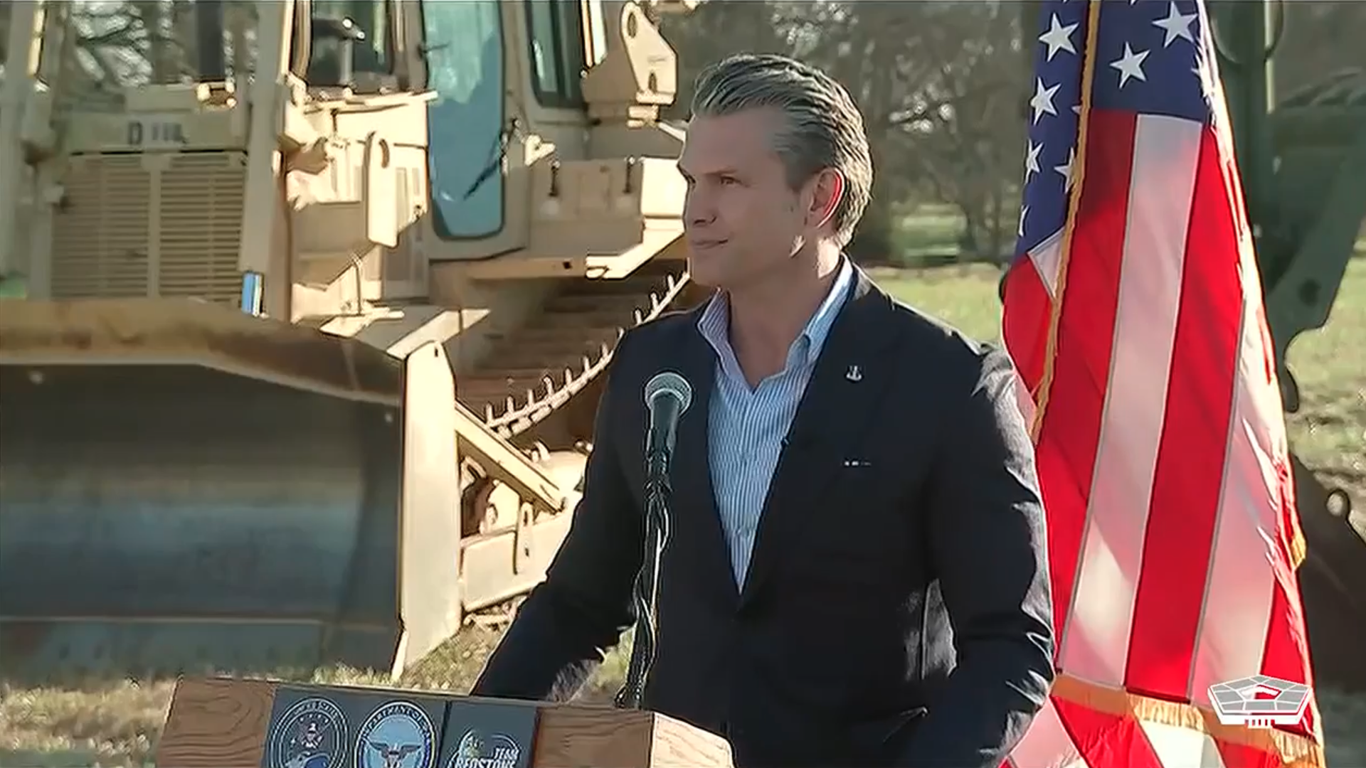 Person in a dark blazer and light blue shirt standing at a podium with seals, microphone, American flag, and construction vehicle in background outdoors.