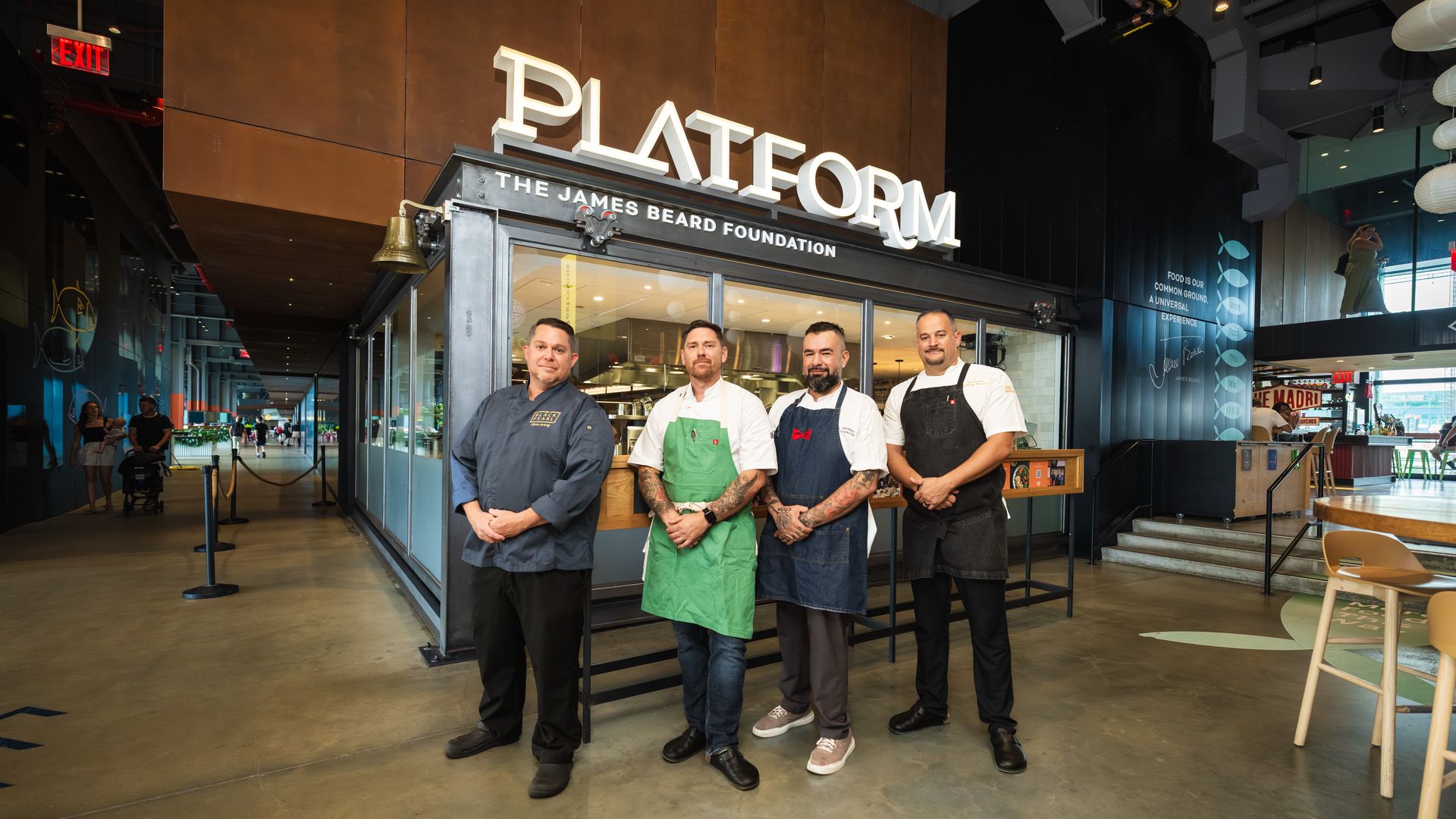Four chefs in aprons standing inside a modern dining area in front of a restaurant labeled PLATFORM by The James Beard Foundation, with seating and a hallway in the background.