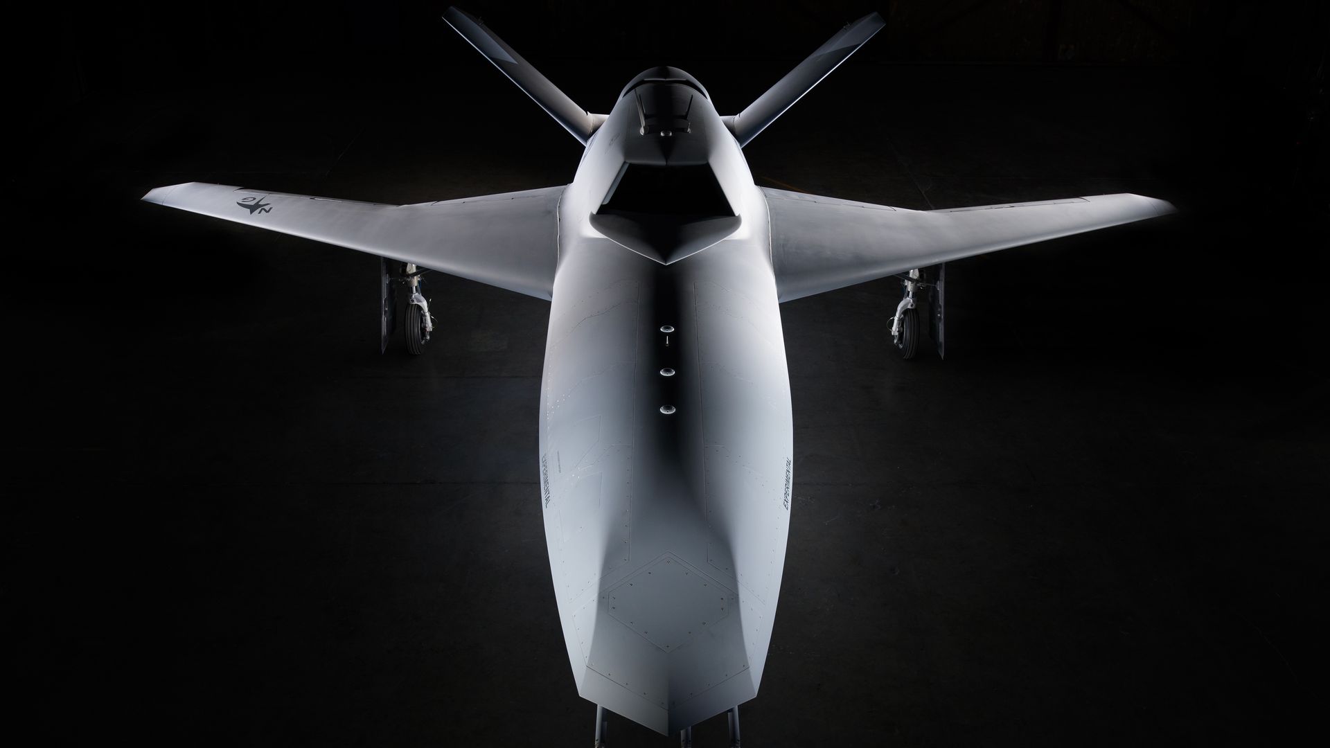 Front view of a sleek, grey drone with angled wings and twin tail fins, positioned in a dark hangar with focused lighting highlighting its aerodynamic design.