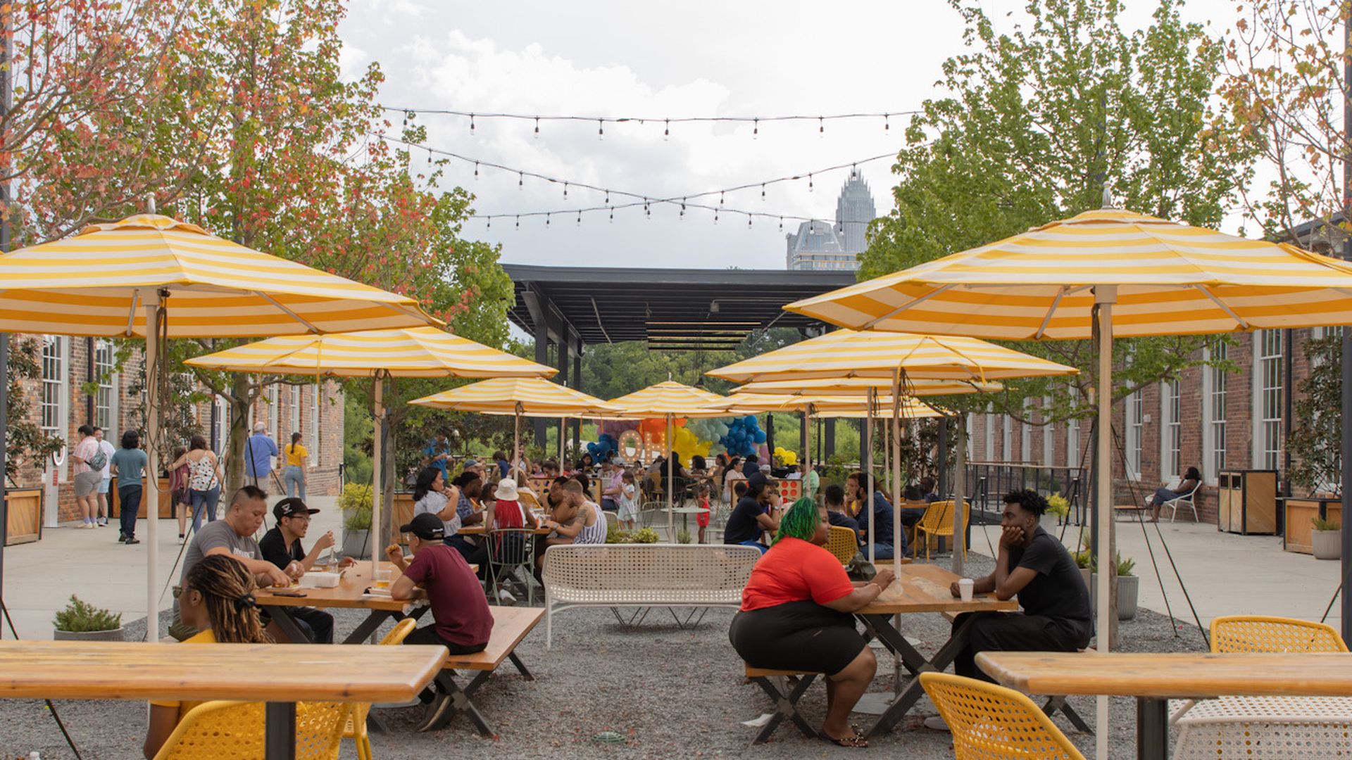 Optimist Hall. Outdoor courtyard with yellow striped umbrellas over wooden picnic tables. People talk and eat beneath string lights; brick buildings line the sides, trees show autumn colors.