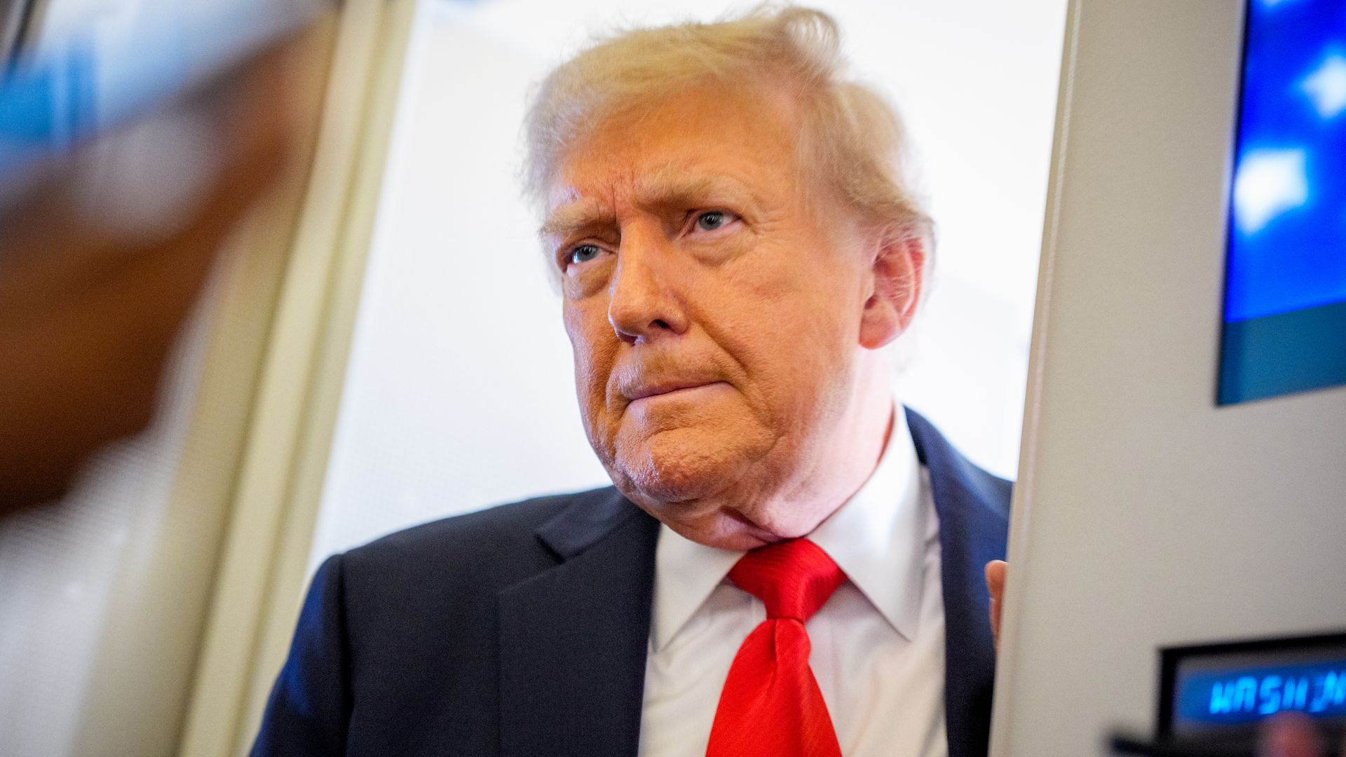 Man with blond hair wearing a black suit, white shirt, and red tie standing indoors near a door with a serious expression.