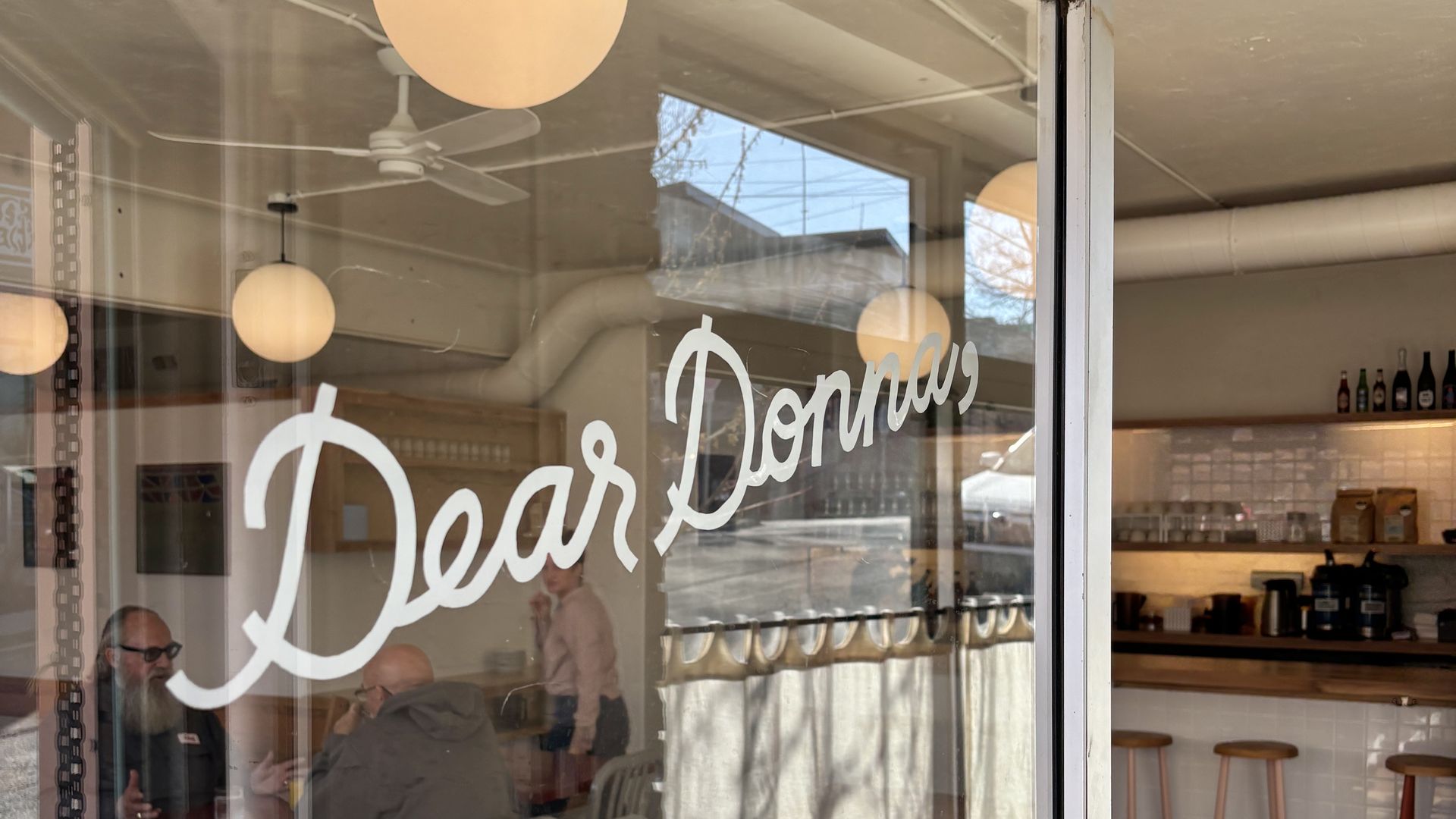 Glass storefront window with white cursive text 'Dear Donnas'; warm cafe interior seen through glass: round pendant lights, a ceiling fan, a counter with stools, bottles on a shelf, and people inside.