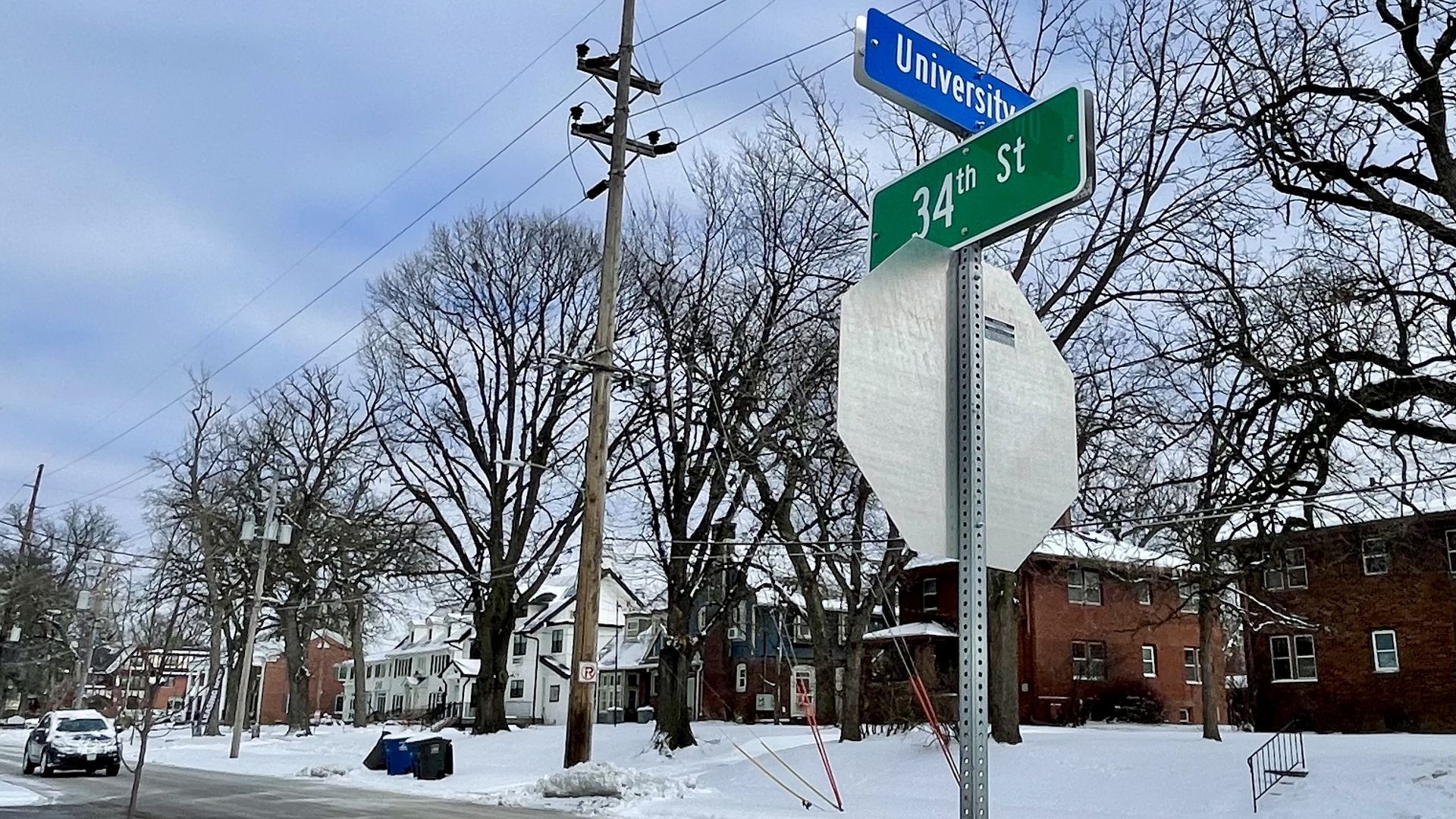 Snow-covered neighborhood street intersection with signs for University and 34th St, leafless trees, parked car, and overcast sky in winter.