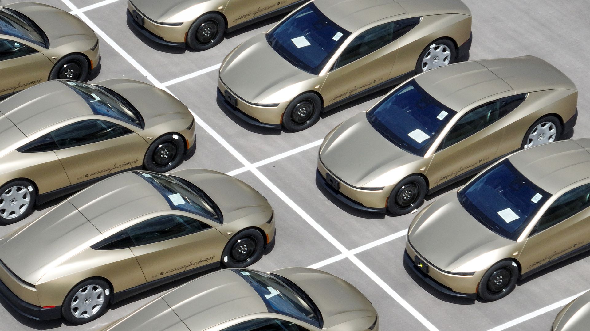 An aerial view of about 10 gold Tesla Cybercabs parked nose to nose in a parking lot, with parking lines visible between them.