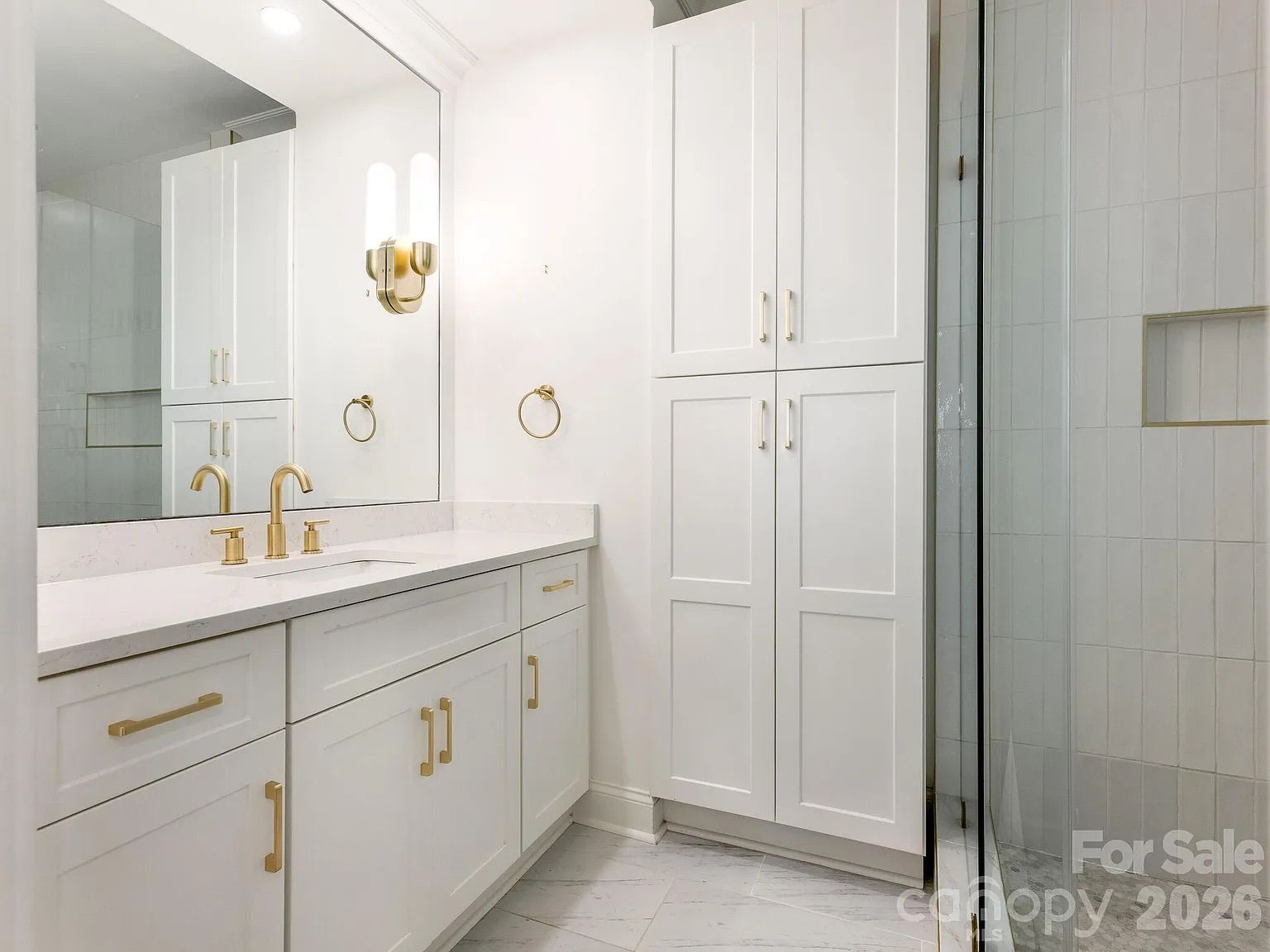 Modern white bathroom with gold fixtures, large mirror, double sink vanity, tall cabinets, and glass shower door with white tile walls and gray floor tiles.