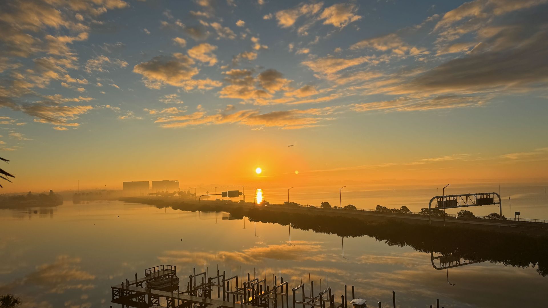 Sunrise over a calm waterway with a dock and boat in the foreground, orange and yellow hues reflecting in the water, scattered clouds in blue sky, and a roadway along the right side.