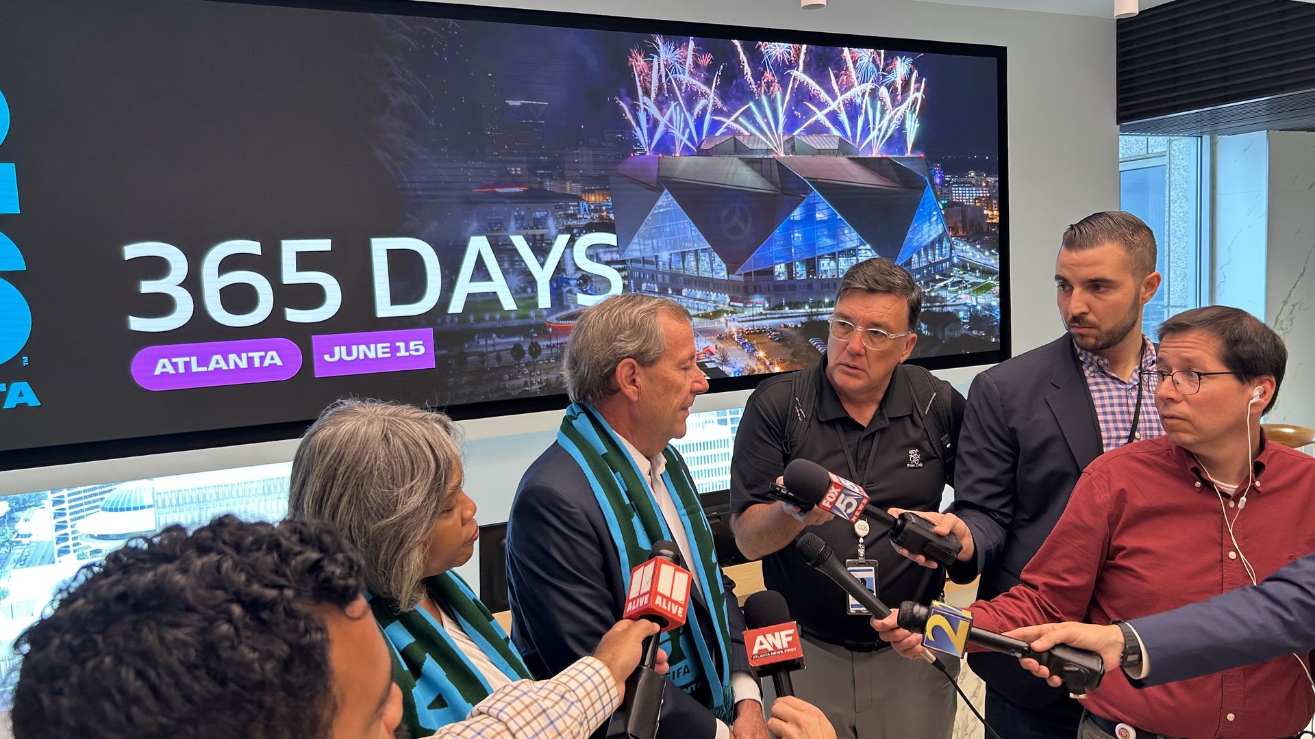 Reporters huddle around two individuals wearing soccer scarves and standing in front of a large digital screen reading "365 DAYS"