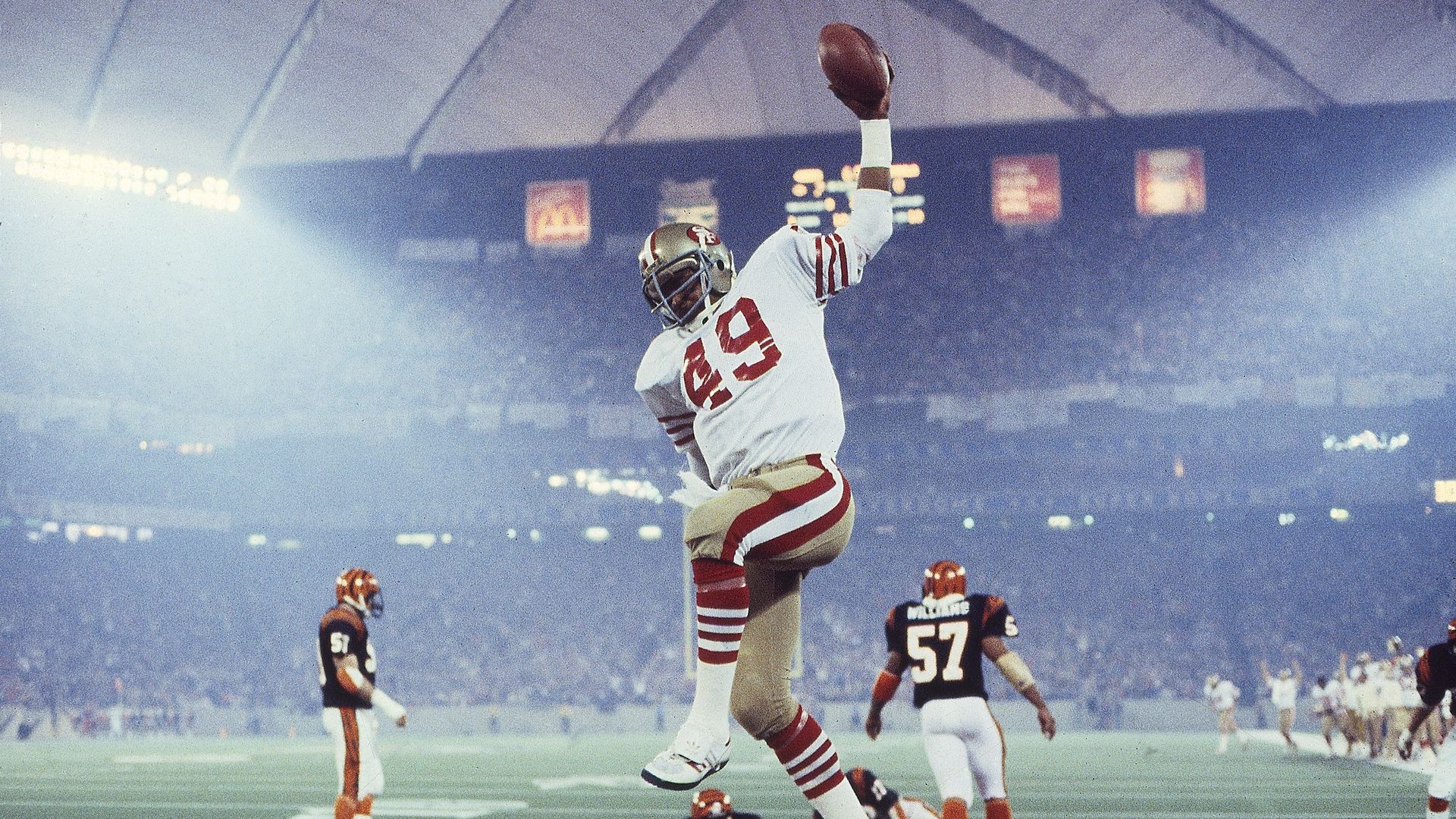 Photo of a 49ers football player leaping in the air with a football in his hand during a touchdown in a packed stadium