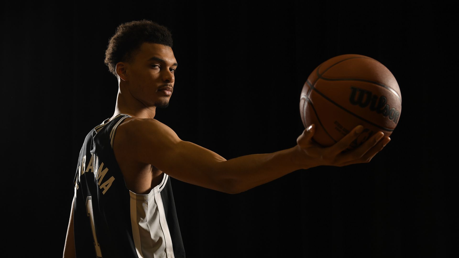Basketball player Victor Wembanyama stands in front of a dark background holding out a basketball with his palm.
