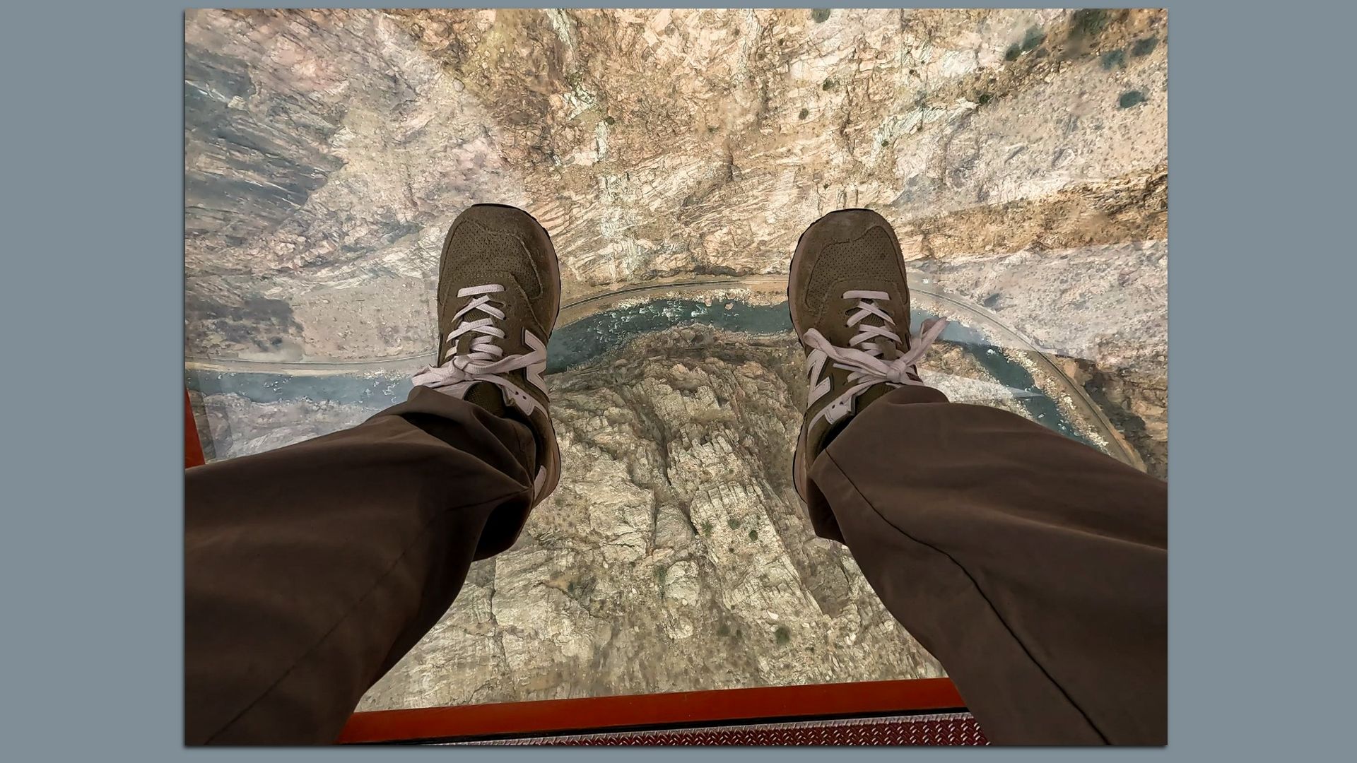 The vew of someone's legs as they look down at their shoes and out the bottom of a glass-bottomed gondola at the Royal Gorge. The view below the gondola is of a steep tan stone canyon with an s-curve of blue river far below. 