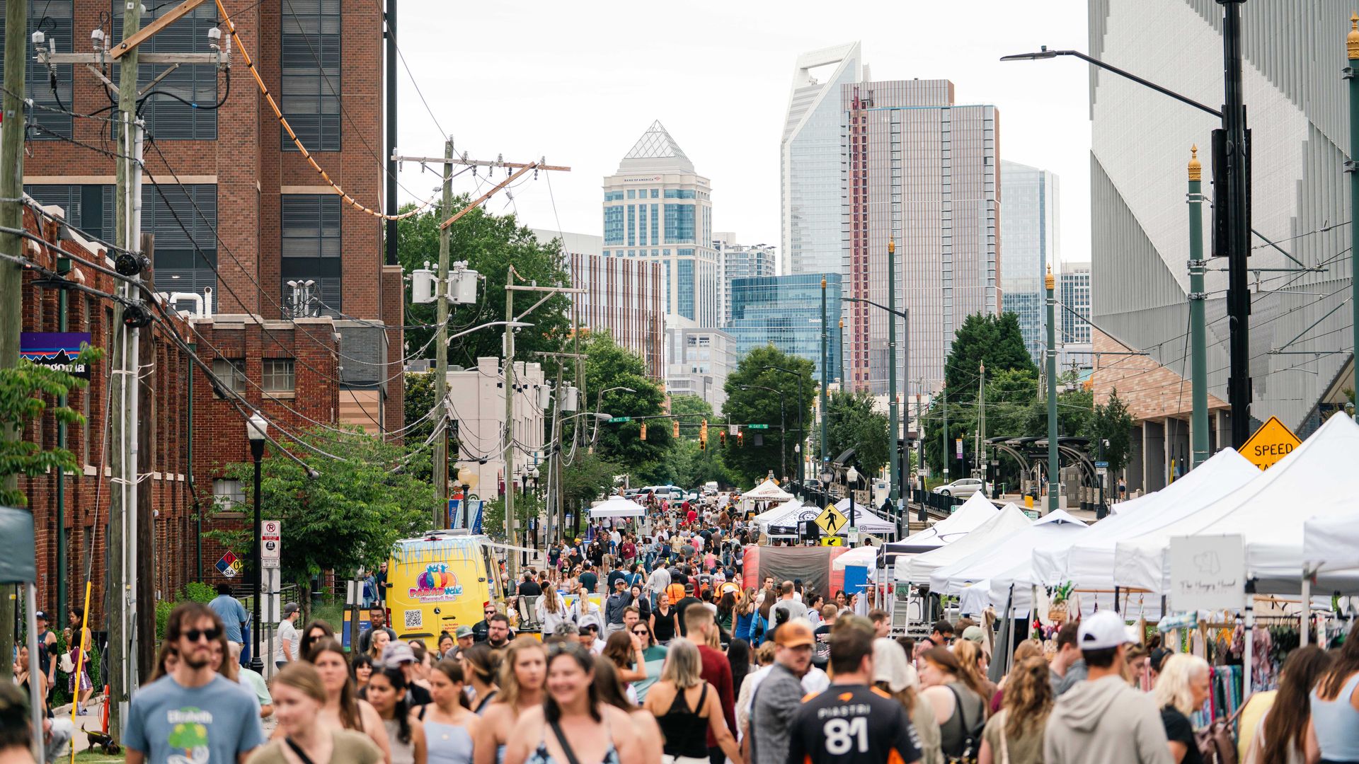 Busy city street festival with white vendor tents lining both sides, a dense crowd of shoppers, and tall modern skyscrapers in the distance behind brick buildings and utility poles.